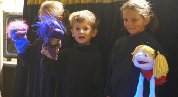 Several children hold puppets in front of a black curtain