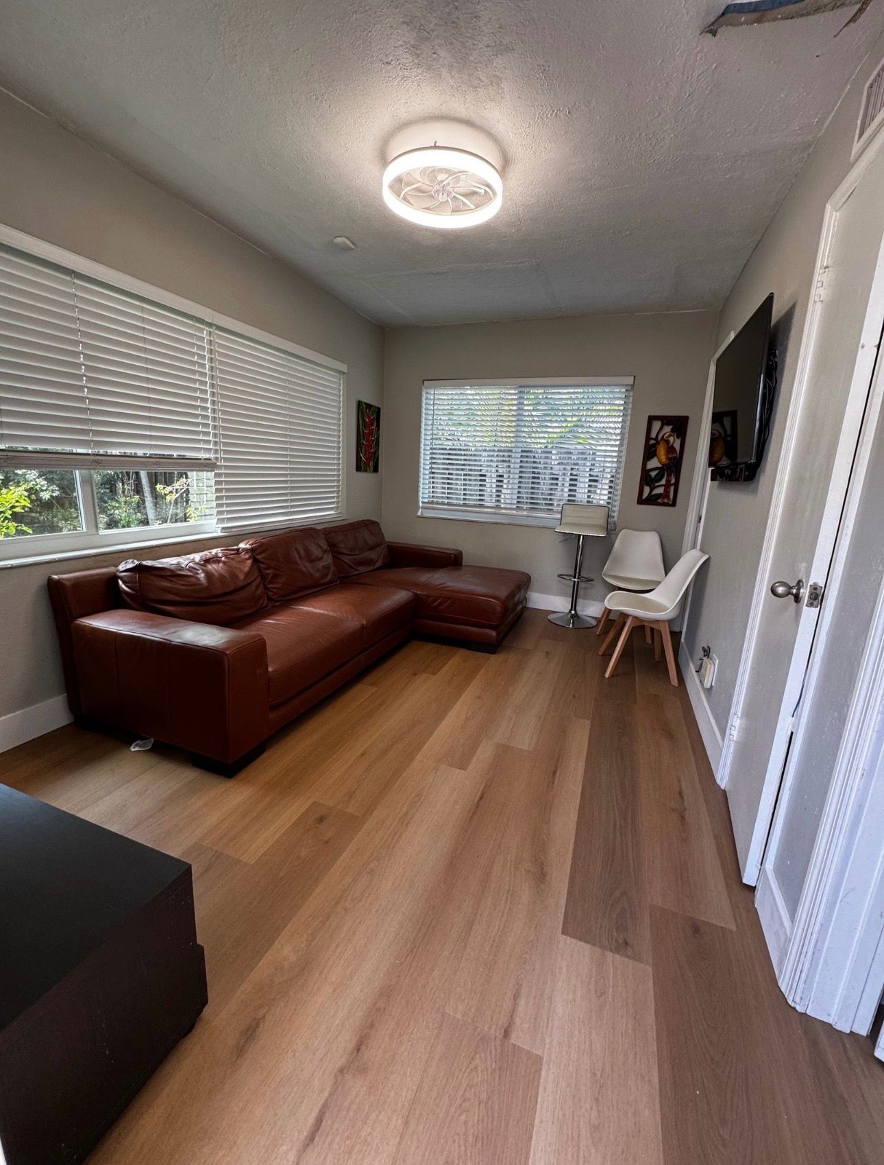 Bright living room with brown leather sectional, wooden floor, large window, and white chairs.