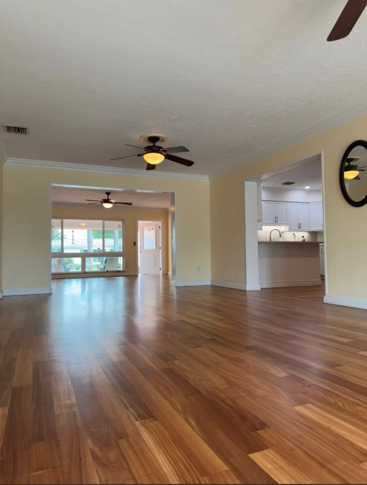 Empty living room with hardwood floors, beige walls, ceiling fans, and a view into the kitchen and dining area.