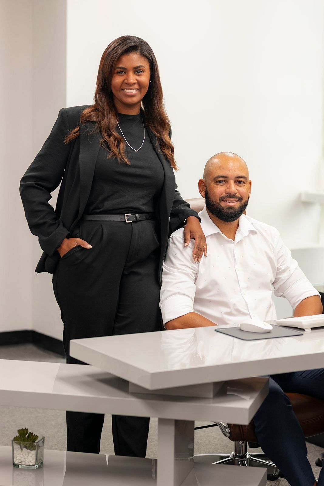 Two colleagues in a meeting room, one standing beside a seated man at a desk.