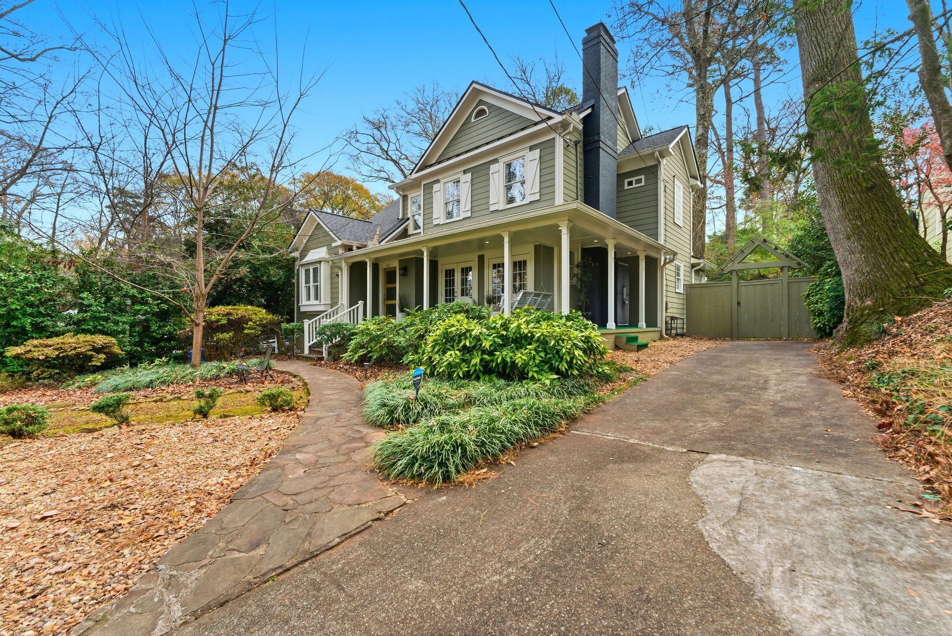 A two-story green house with a covered front porch, situated in a wooded yard with a paved driveway and walkway.