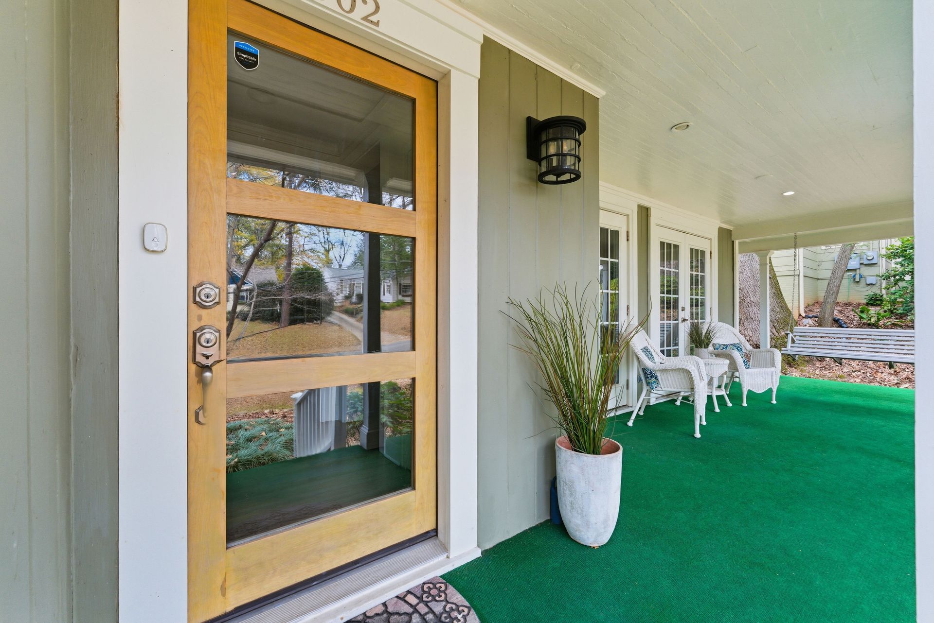 A modern wooden front door with three glass panels on a porch with green flooring, a white planter, and seating.