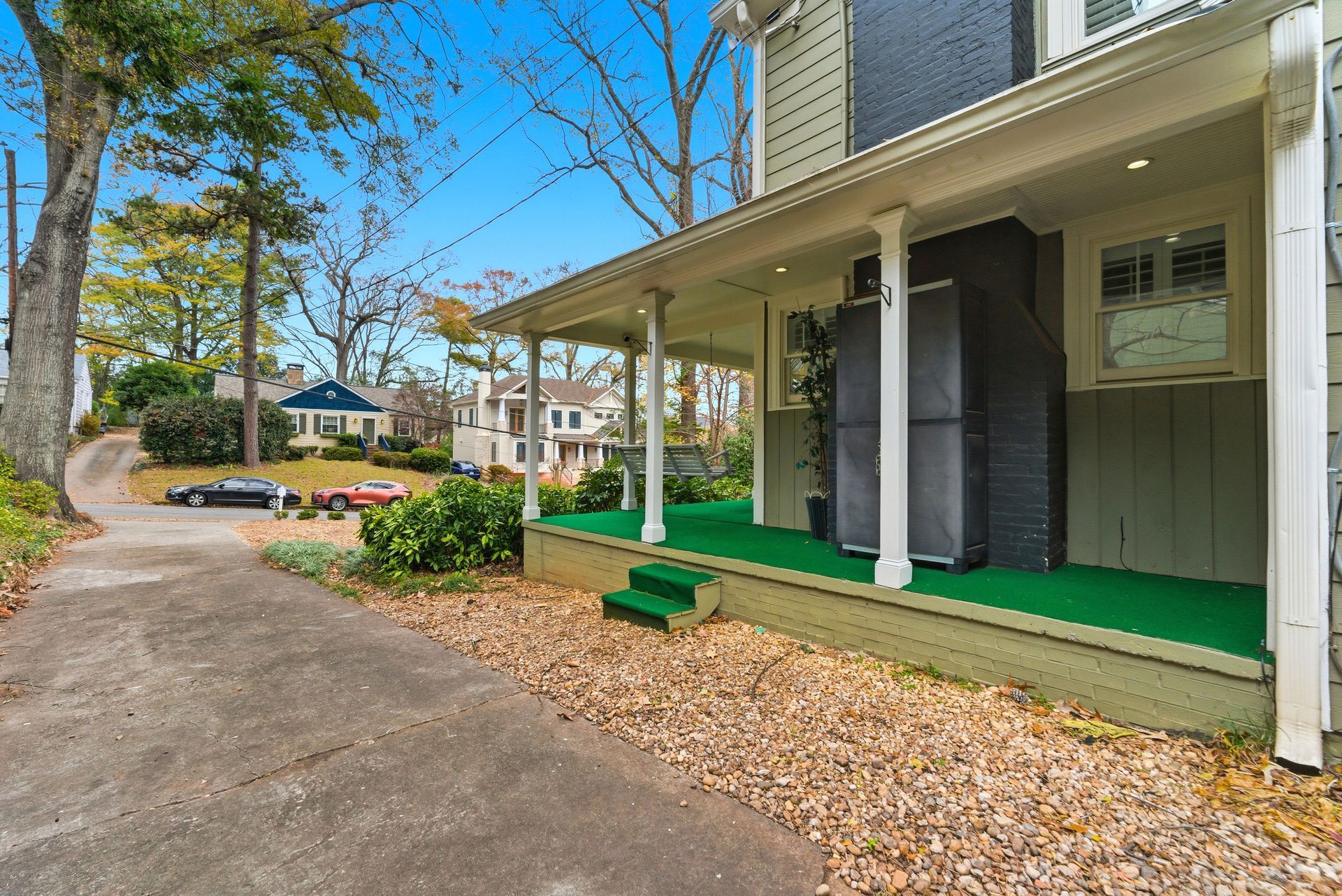 A side view of a house with a covered porch featuring green carpeting, leading to a concrete driveway and trees.