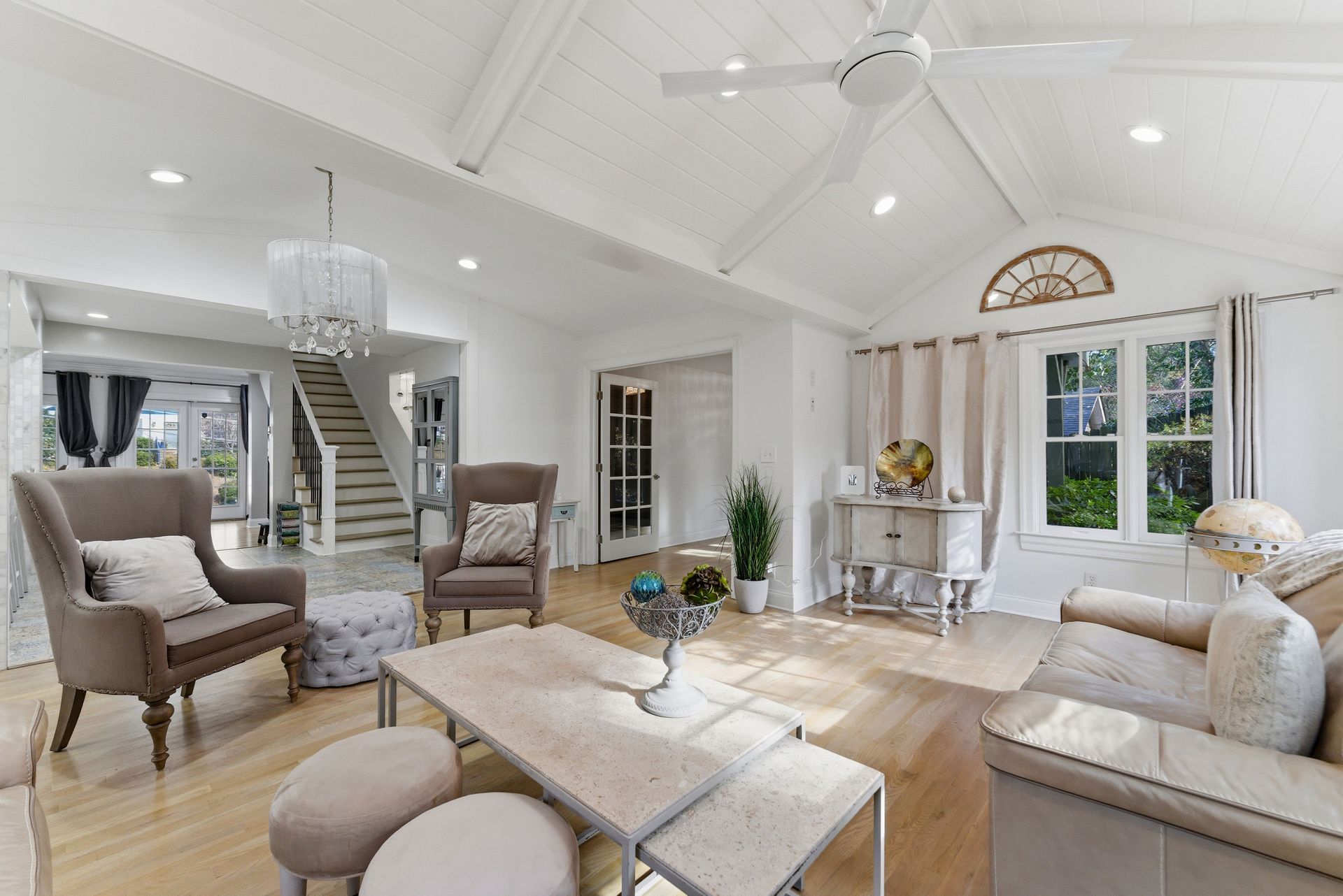 A bright, airy living room with a vaulted ceiling, beige armchair, sofa, ottoman, and wood flooring.
