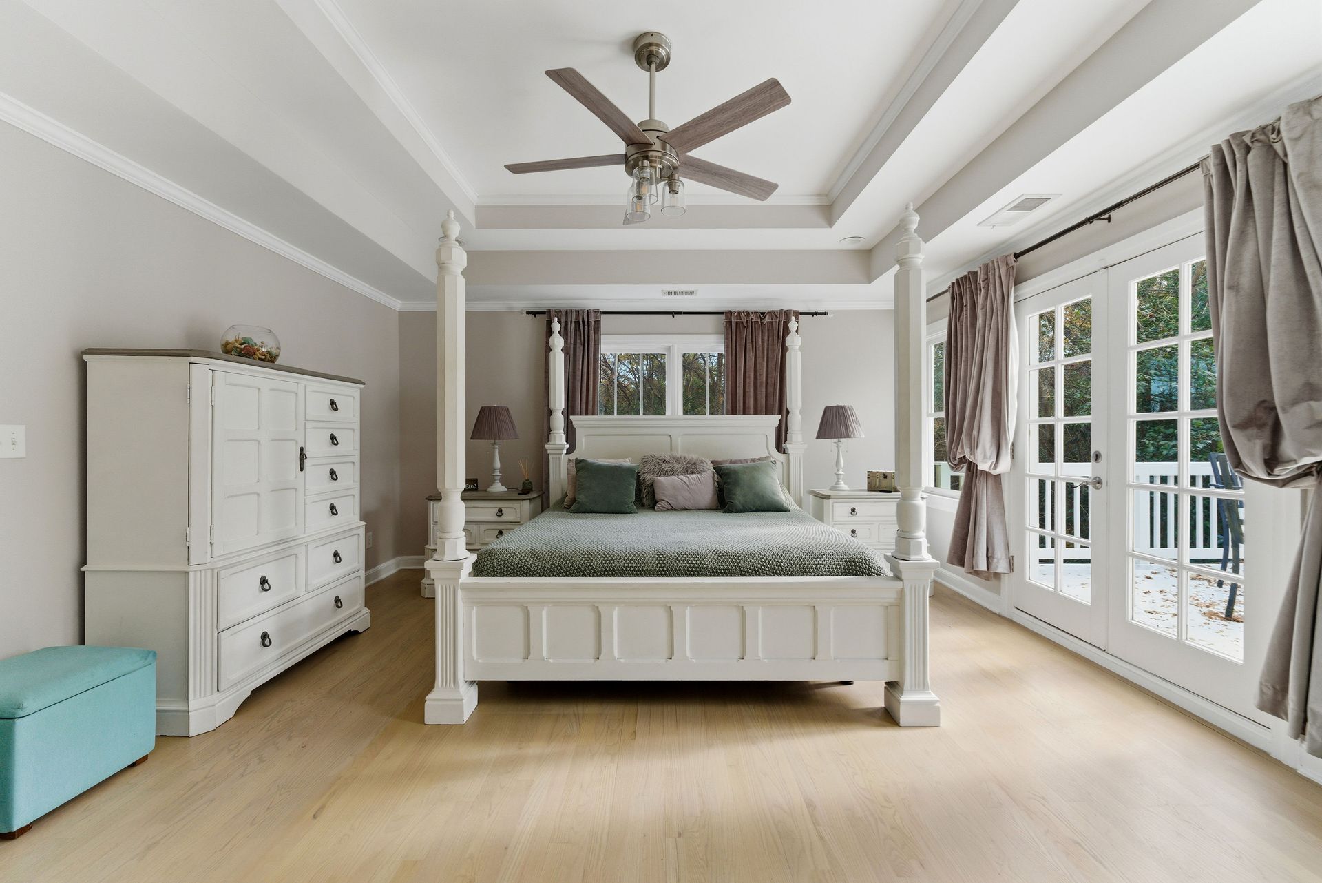 A bright bedroom with a white canopy bed, a tall matching dresser, a light blue ottoman, and glass doors to a balcony.