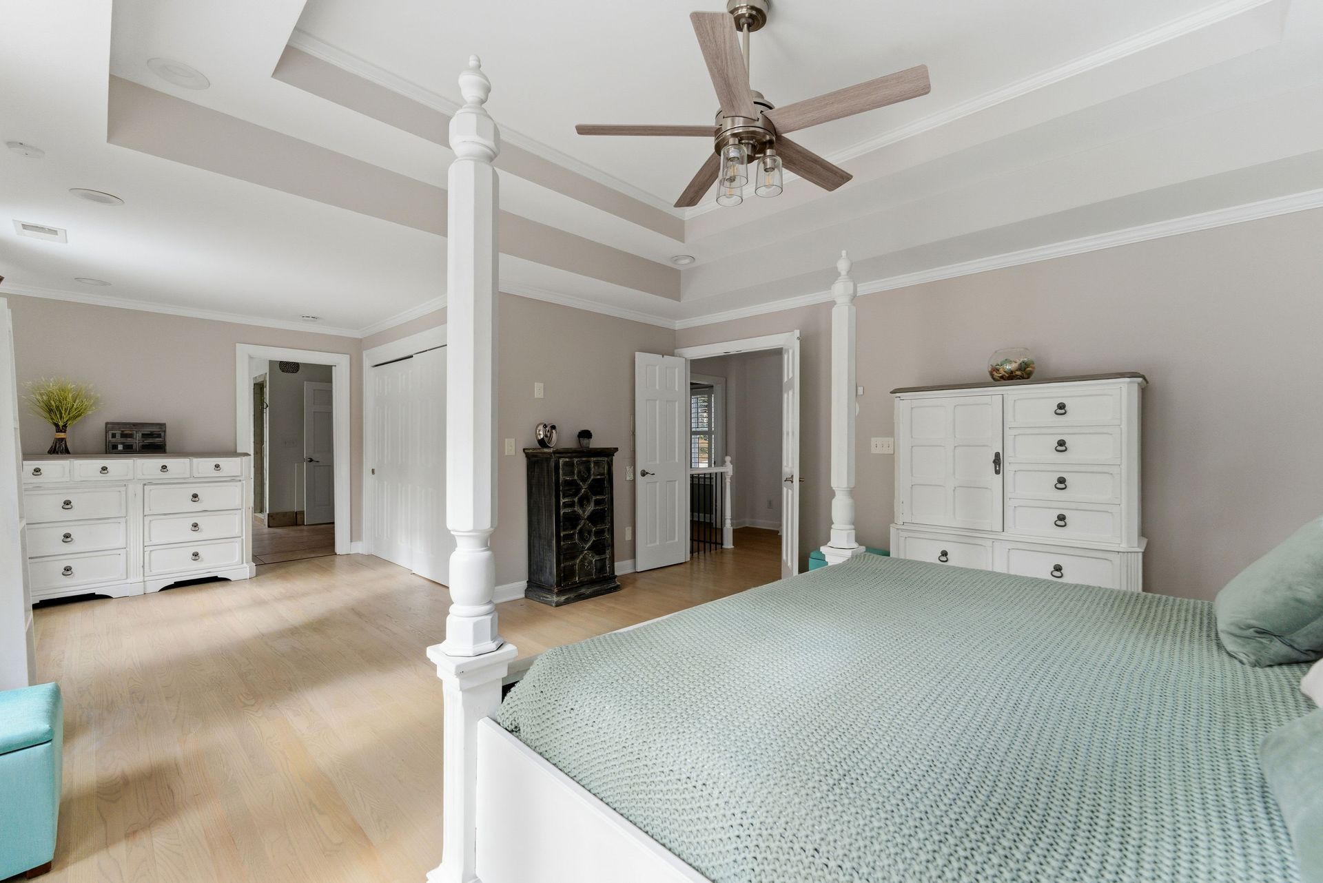 A bright, spacious primary bedroom with a four-poster bed, light wood floors, and white furniture in a neutral room.