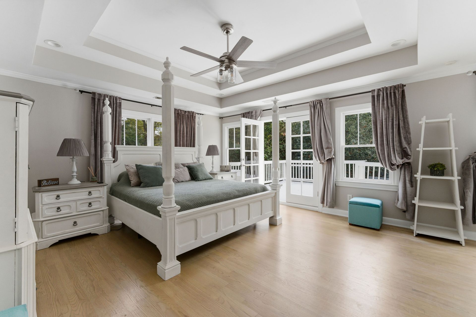 A bright bedroom with a white four-poster bed, wood floors, tray ceiling, and glass doors opening to a balcony.