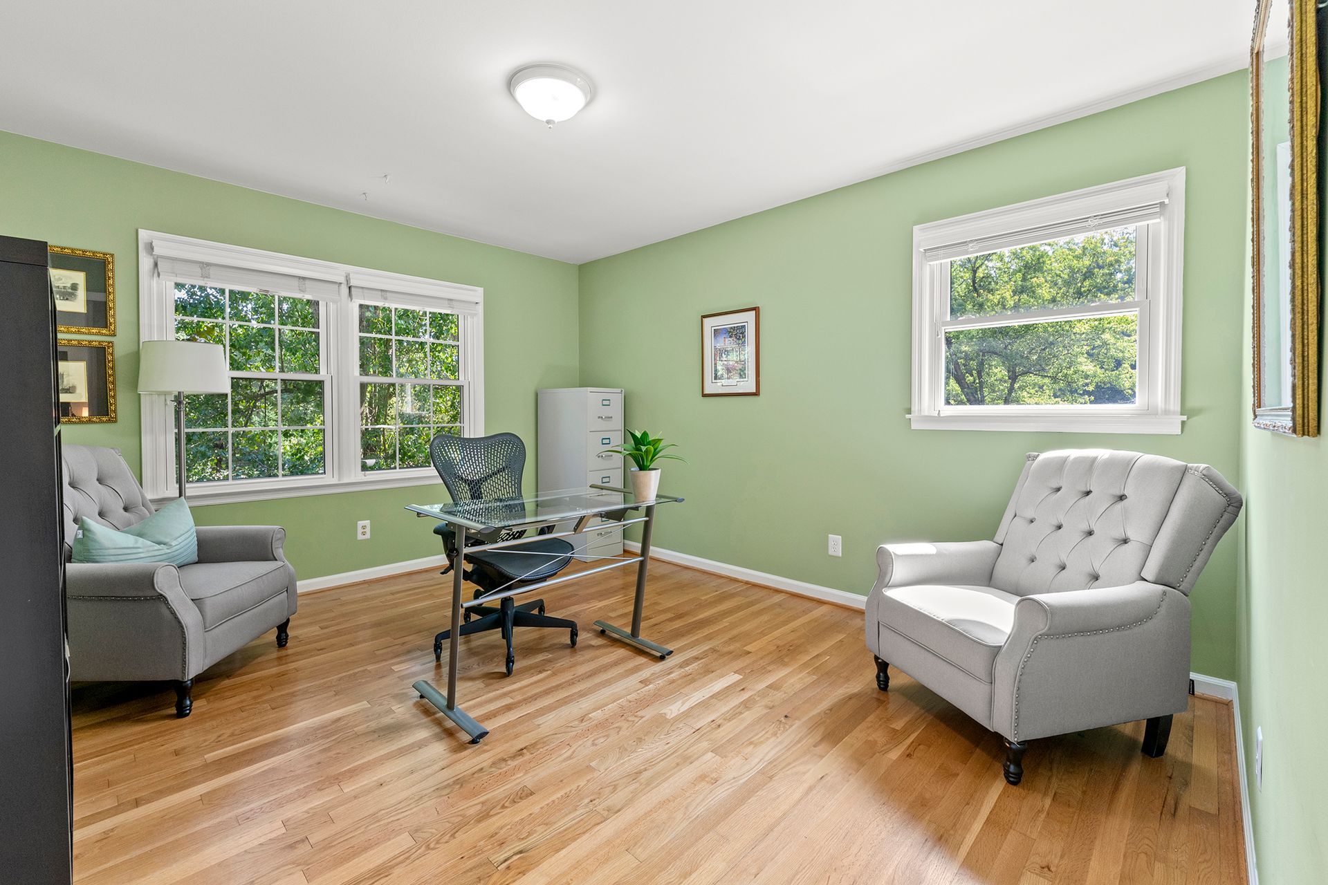 A living room with green walls , hardwood floors , two chairs , a desk and two windows.