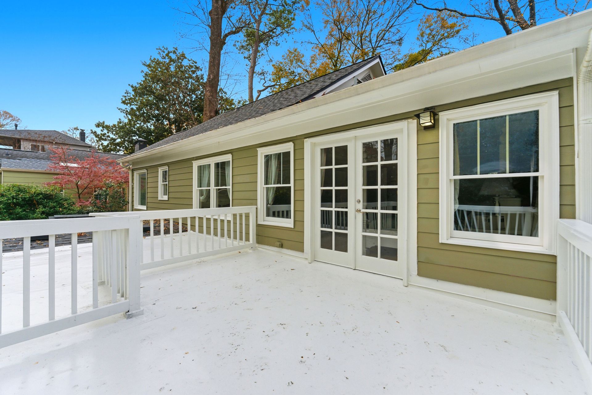 An elevated wooden deck with white railings, featuring sage green siding, French doors, and windows against a blue sky.