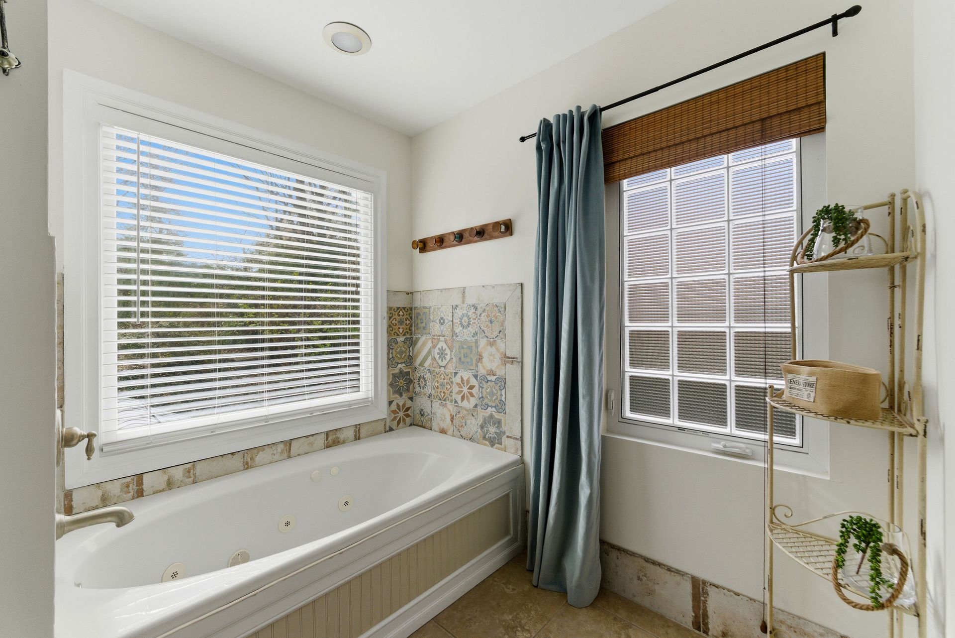 A bathroom with a white soaking tub, decorative tile backsplash, blue curtains, two windows, and a metal shelving unit.
