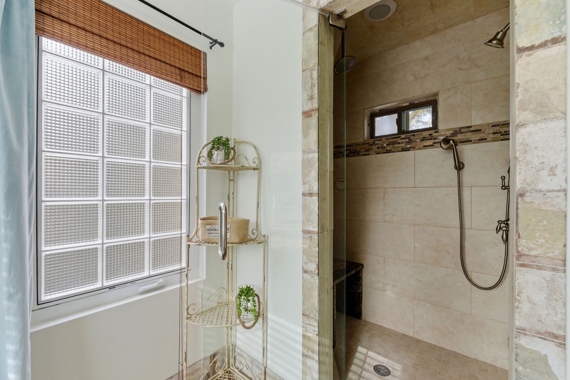 A walk-in shower with stone tile walls, glass door, and a nearby metal shelf under a block window with a woven shade.