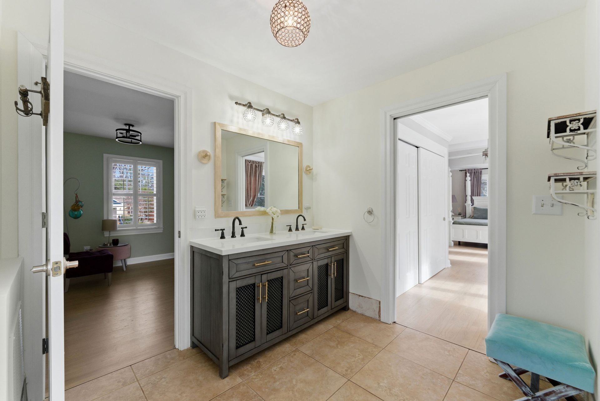 A bathroom with a dark gray dual-sink vanity, light beige tile floors, and doorways leading to adjacent rooms.