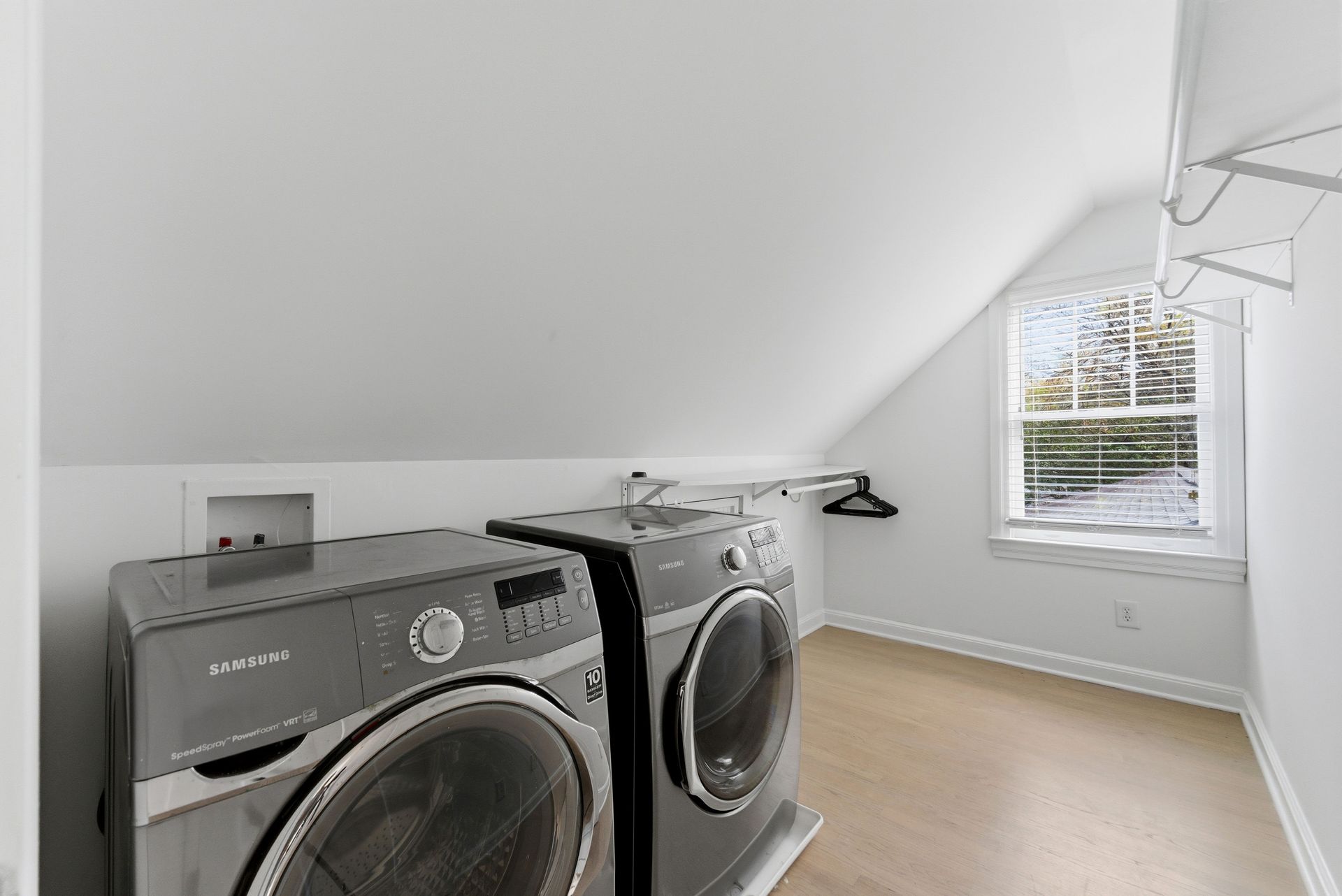 A laundry room with two dark gray, front-loading appliances, white slanted walls, a window, and a wall-mounted clothes rod.