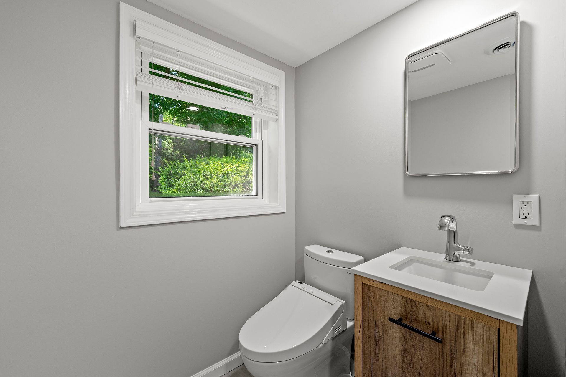 A bathroom with a toilet , sink , mirror and window.