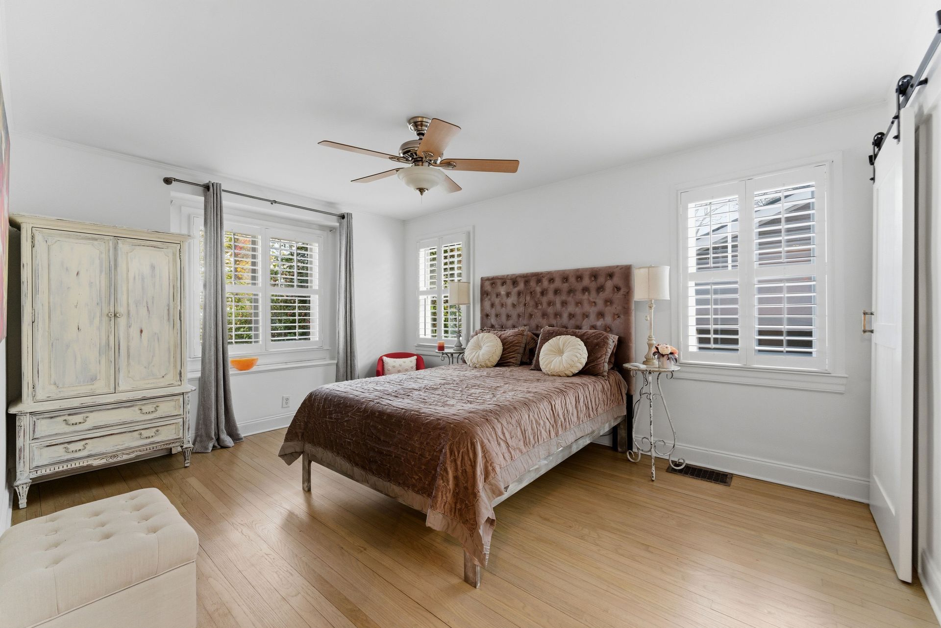 A bright bedroom with hardwood floors, a tufted headboard, a white dresser, and windows with shutters.