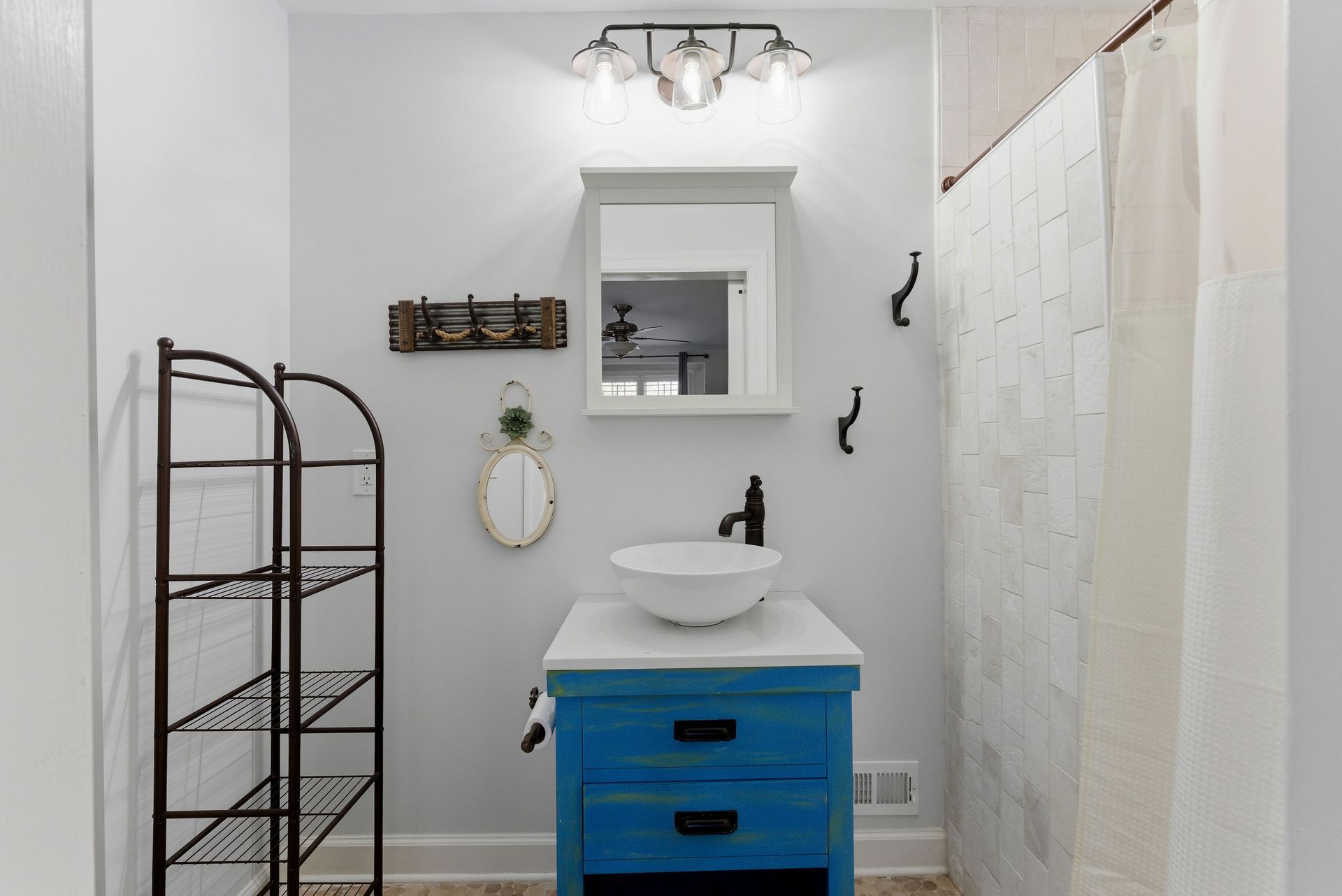 A bathroom with light gray walls, a blue vanity, a vessel sink, a white medicine cabinet, and a metal shelving unit.