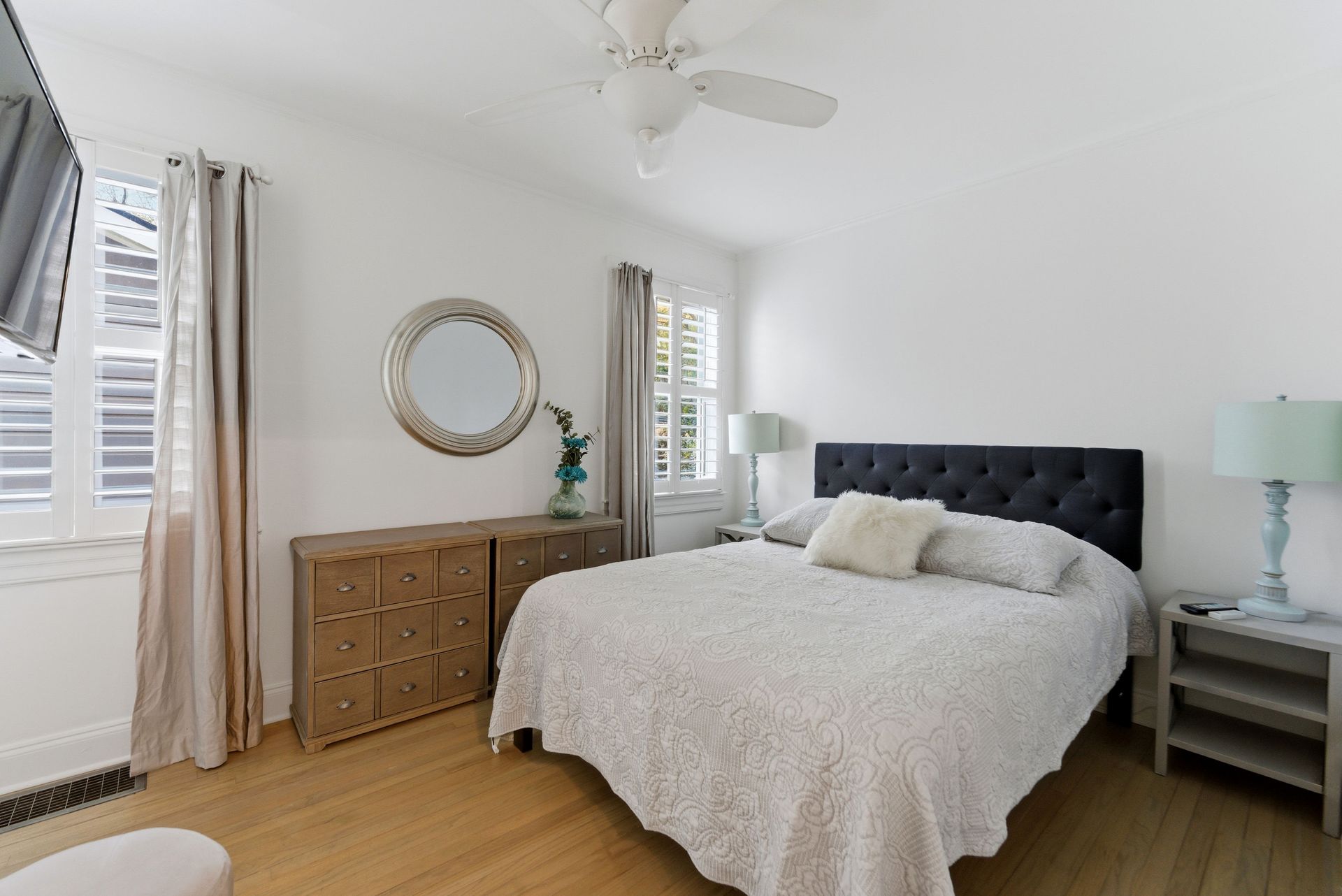 A bedroom with a queen bed, dark headboard, wood dresser, round mirror, and white walls.