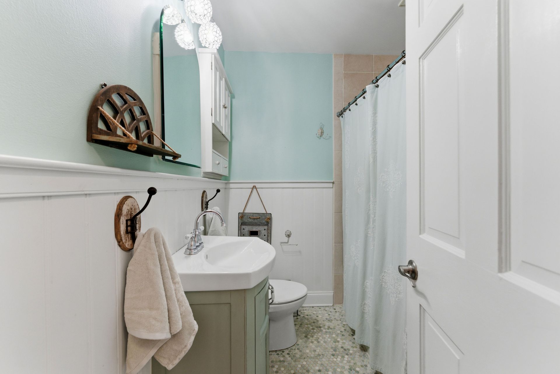 Small bathroom with a sage green vanity, white wainscoting, light blue walls, tiled floor, and a white shower curtain.