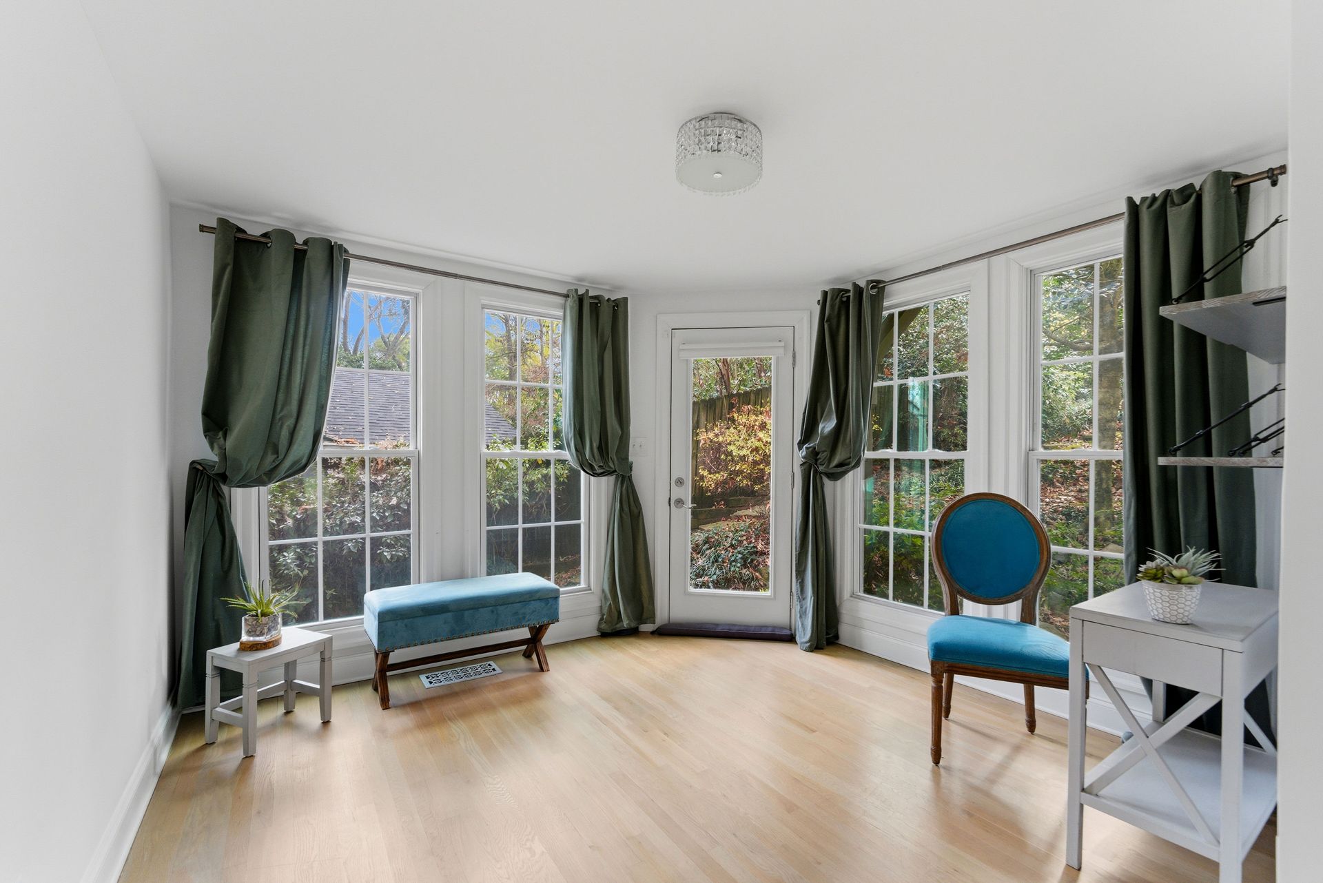 A sunlit room with hardwood floors, a turquoise bench and chair, and tall windows framed by olive green curtains.