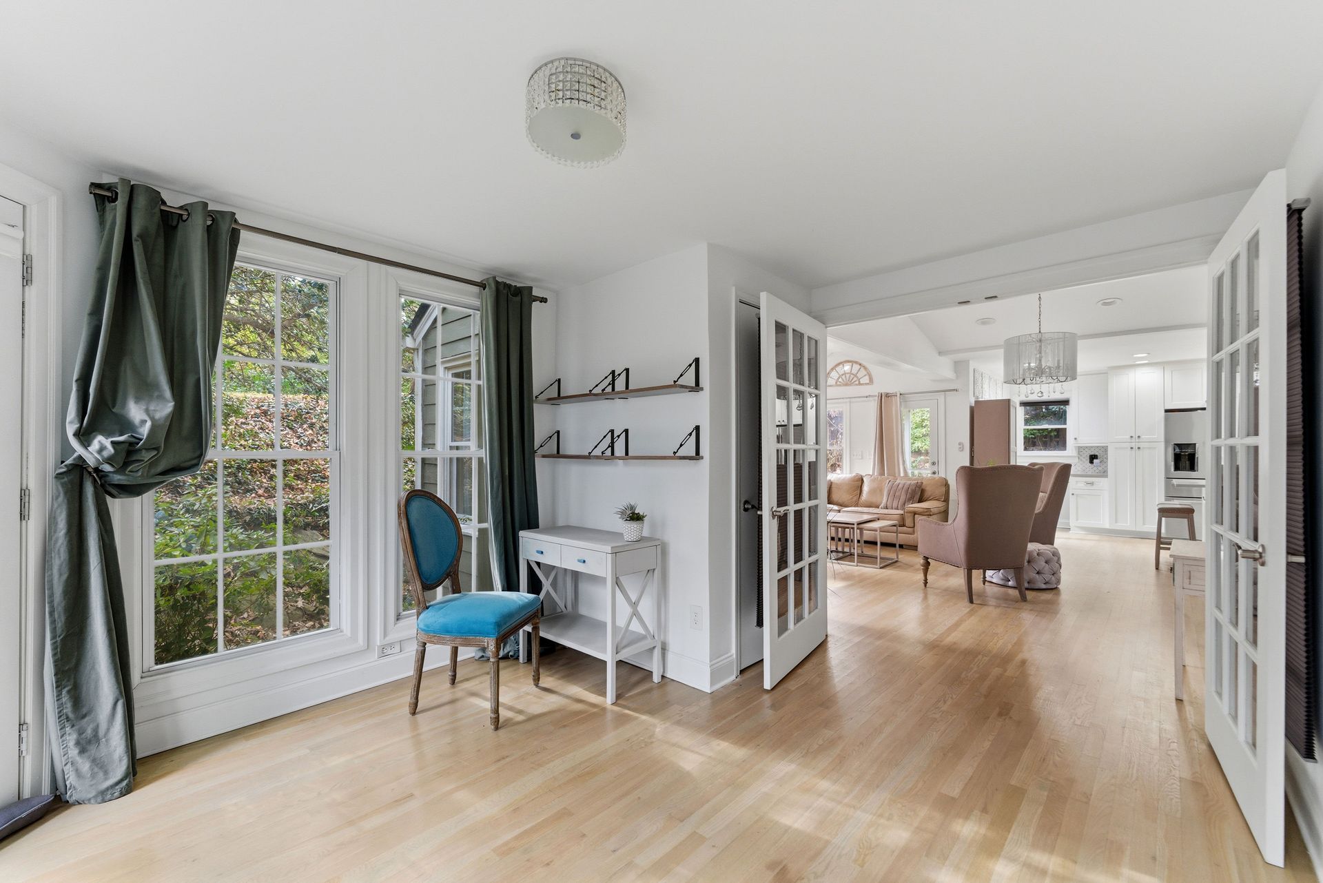 A room with hardwood floors, a teal chair at a small desk, two windows with curtains, and French doors leading to a lounge.