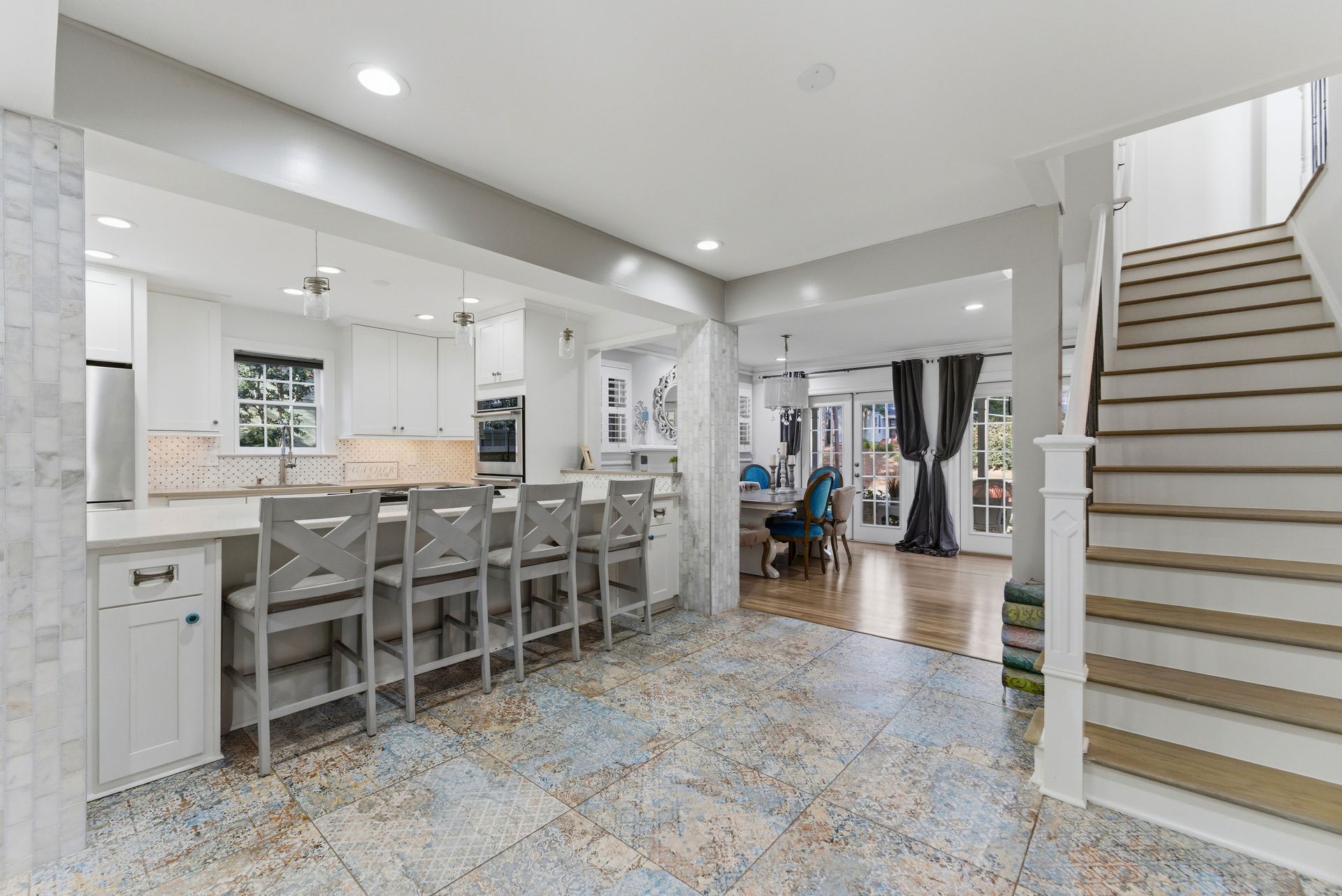 A bright kitchen with a marble-topped island, four gray stools, and a wooden staircase leading to an upper floor.