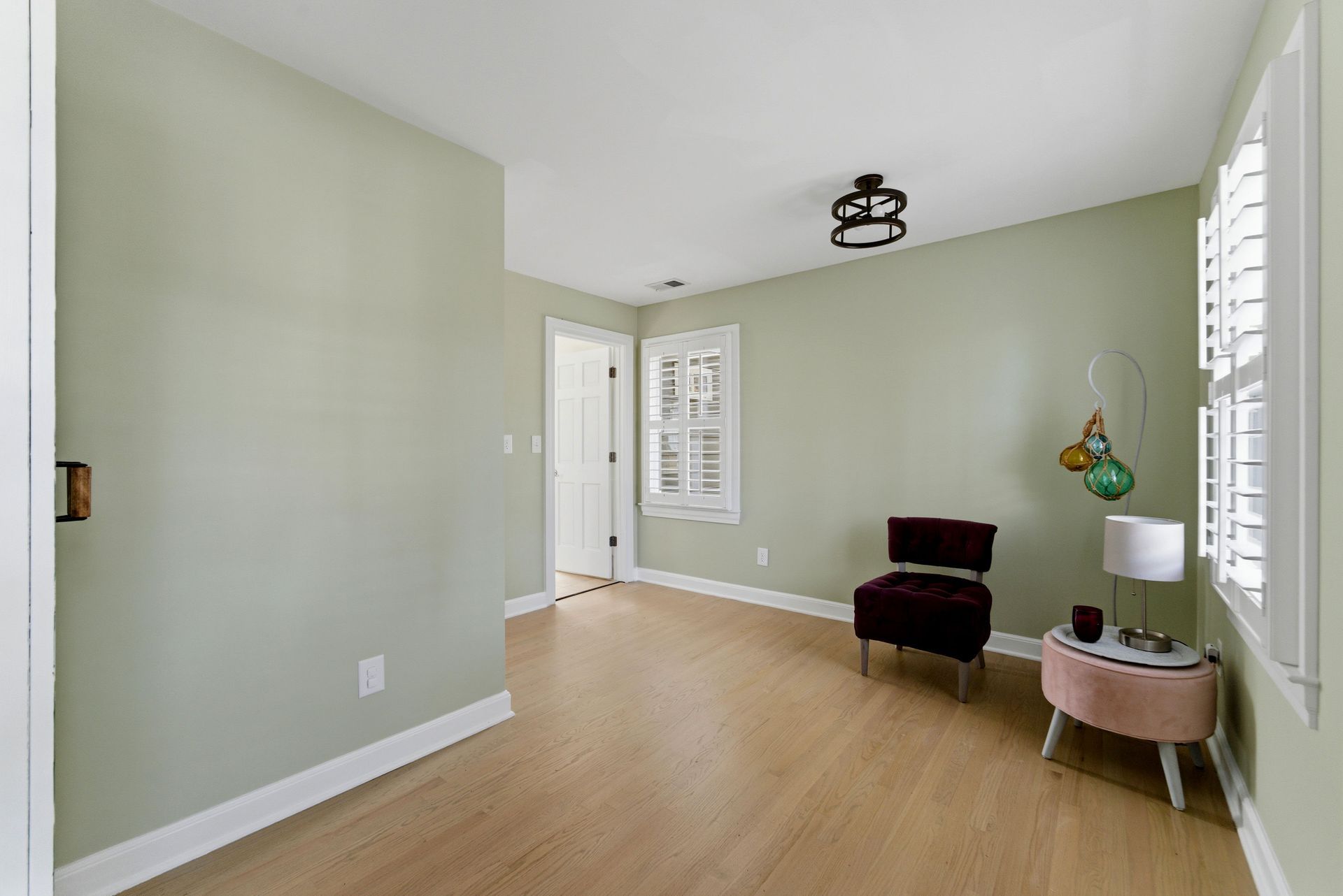 A light sage-green room with wood-look flooring, a dark velvet chair, a side table with a lamp, and white shutter windows.