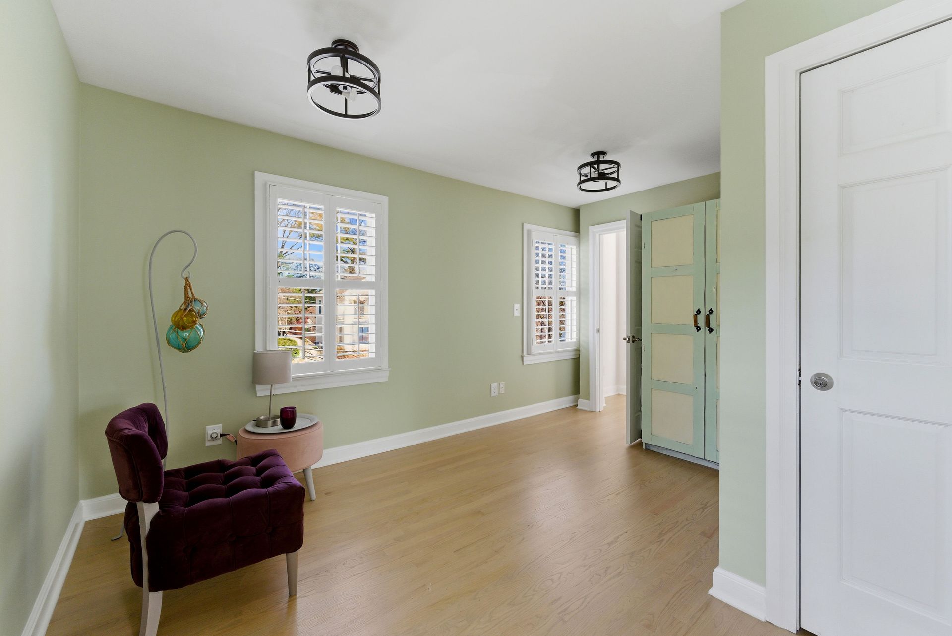 A sunlit, sage green room with light wood floors, a dark purple armchair, a small side table, and two white windows.