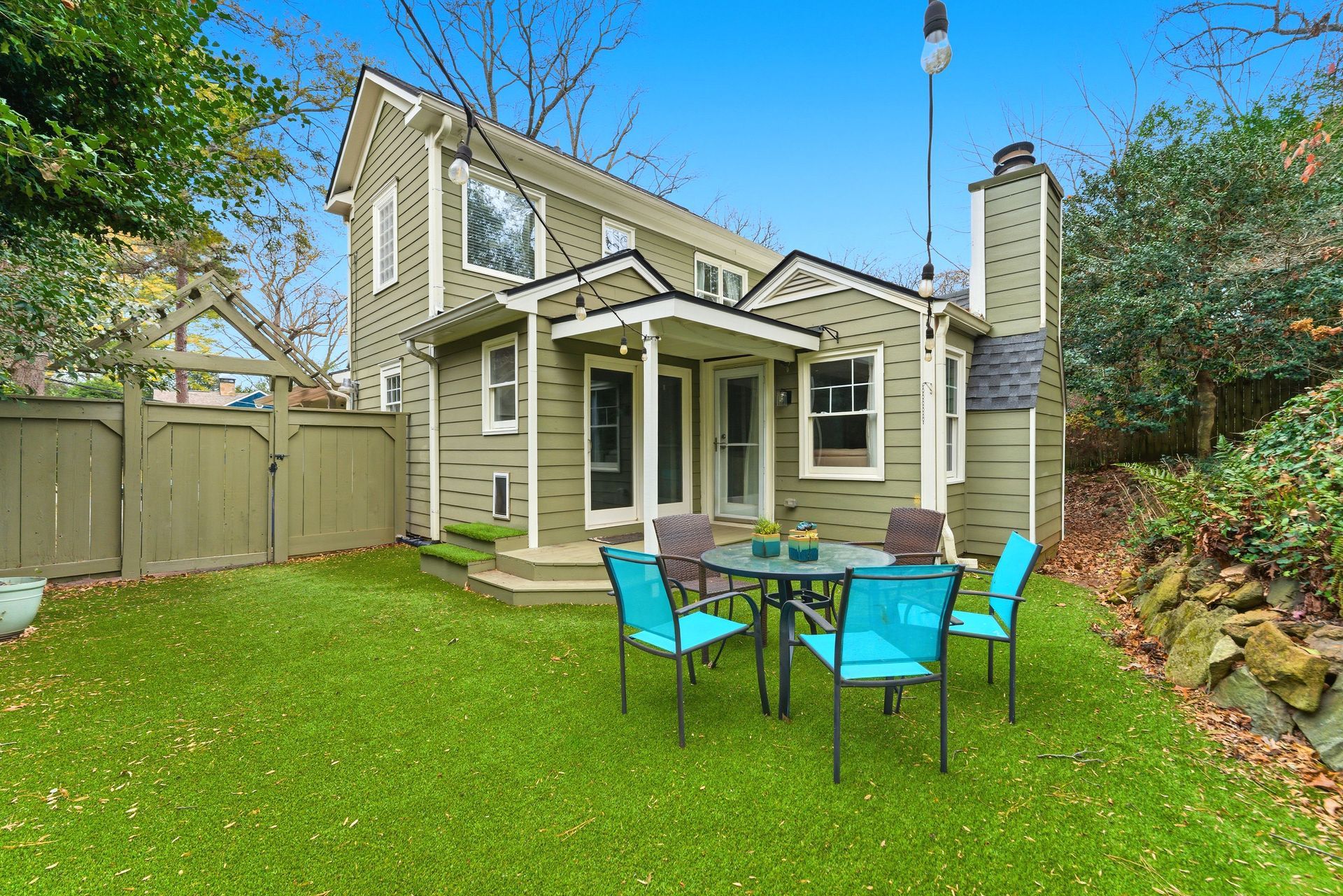 A green two-story house with a small patio, outdoor dining set, and grassy backyard enclosed by a wooden fence.