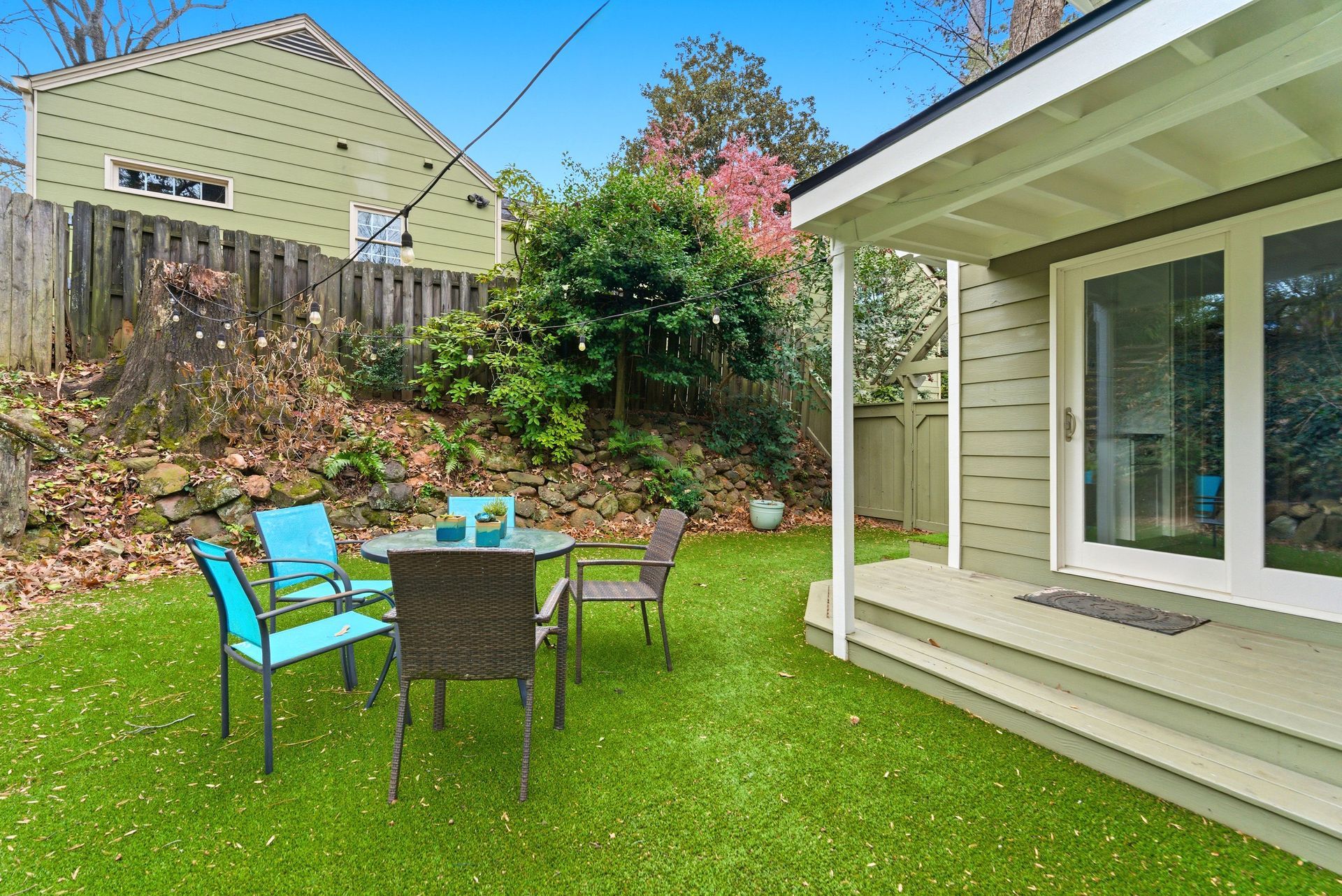 A green lawn with a patio set in front of a house deck, set against a terraced, leafy backyard.