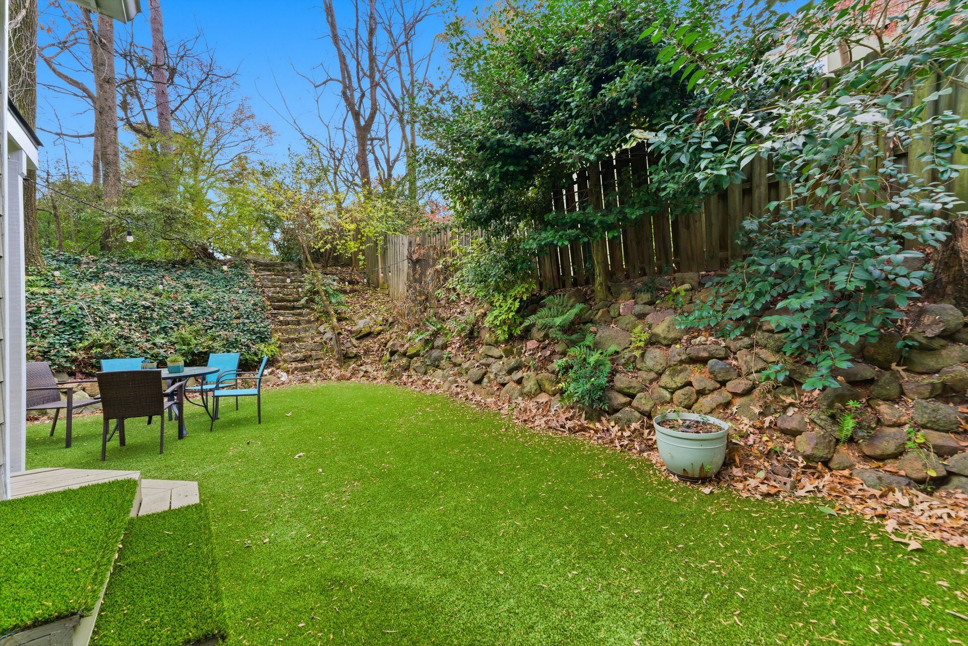 A small backyard with a vibrant green lawn, a stone retaining wall, leafy trees, and patio furniture on the side.