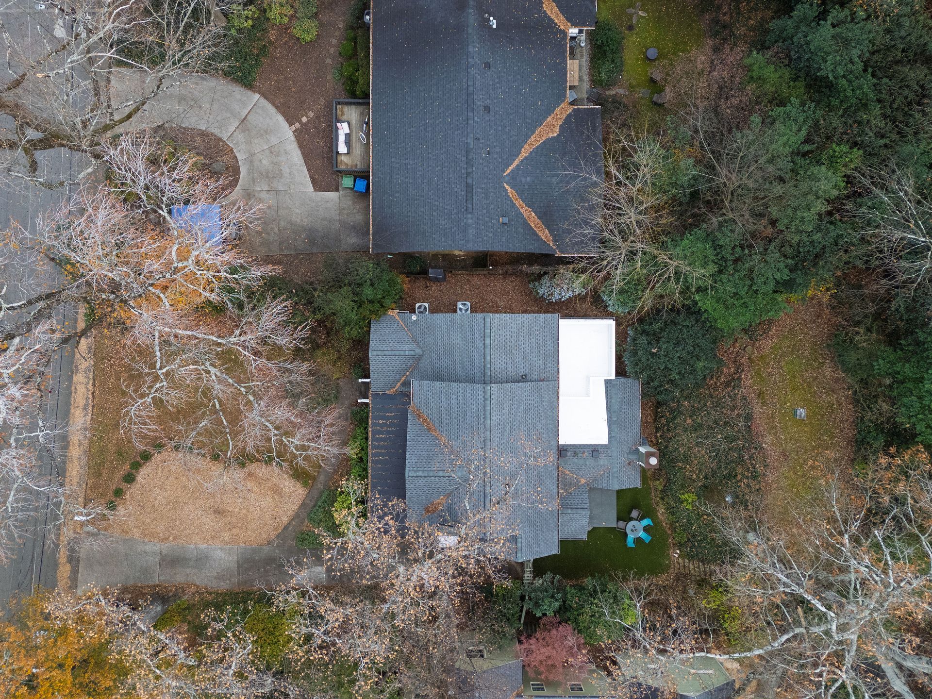 An aerial view of two neighboring residential houses with dark roofs, surrounded by trees and a paved driveway.