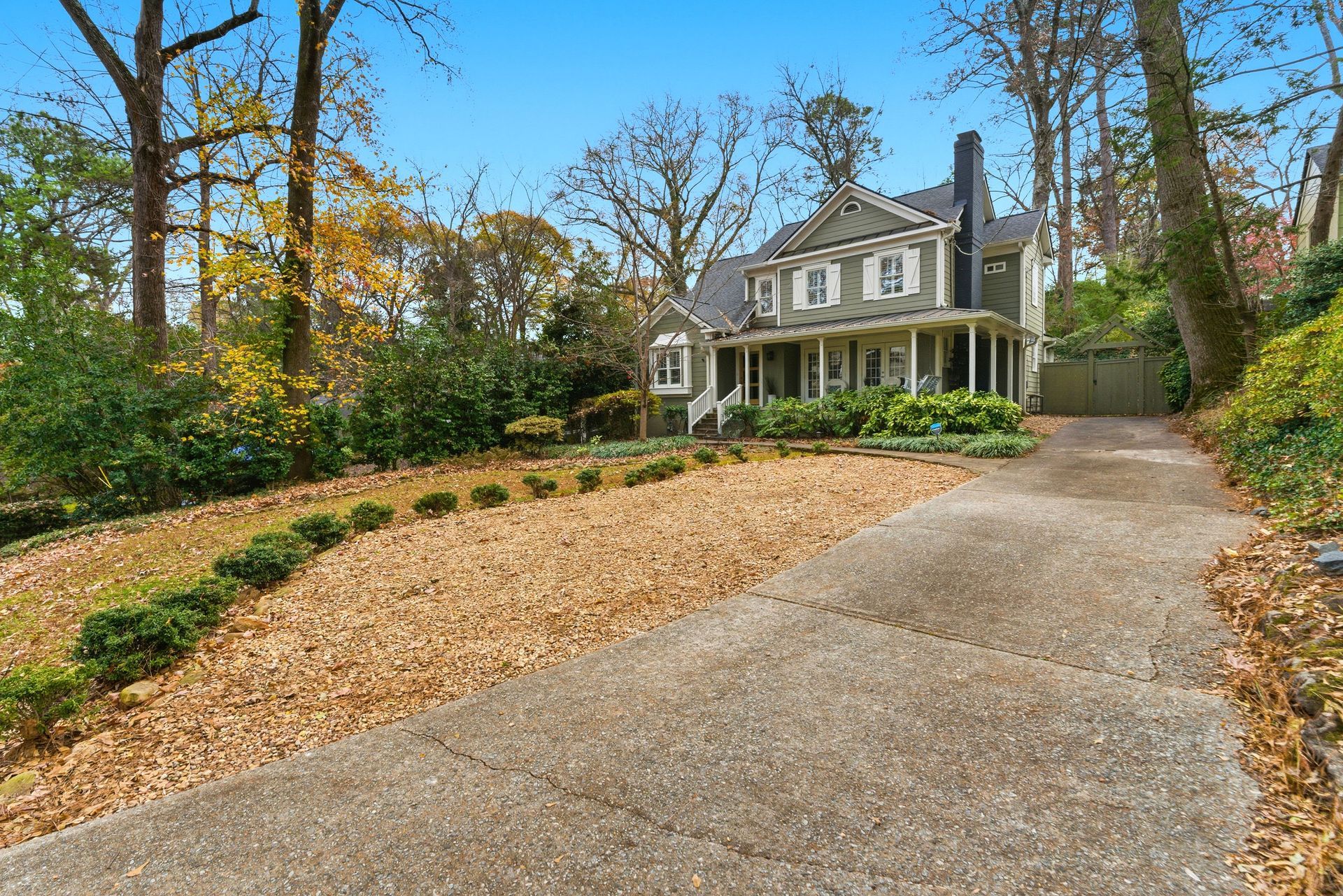 A beige two-story house with a front porch and a prominent chimney, viewed from a long concrete driveway.