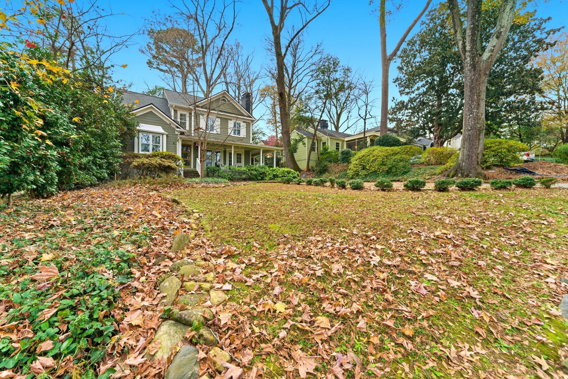 A two-story house with a front porch sits behind a sloping lawn covered in fallen brown leaves under a clear blue sky.