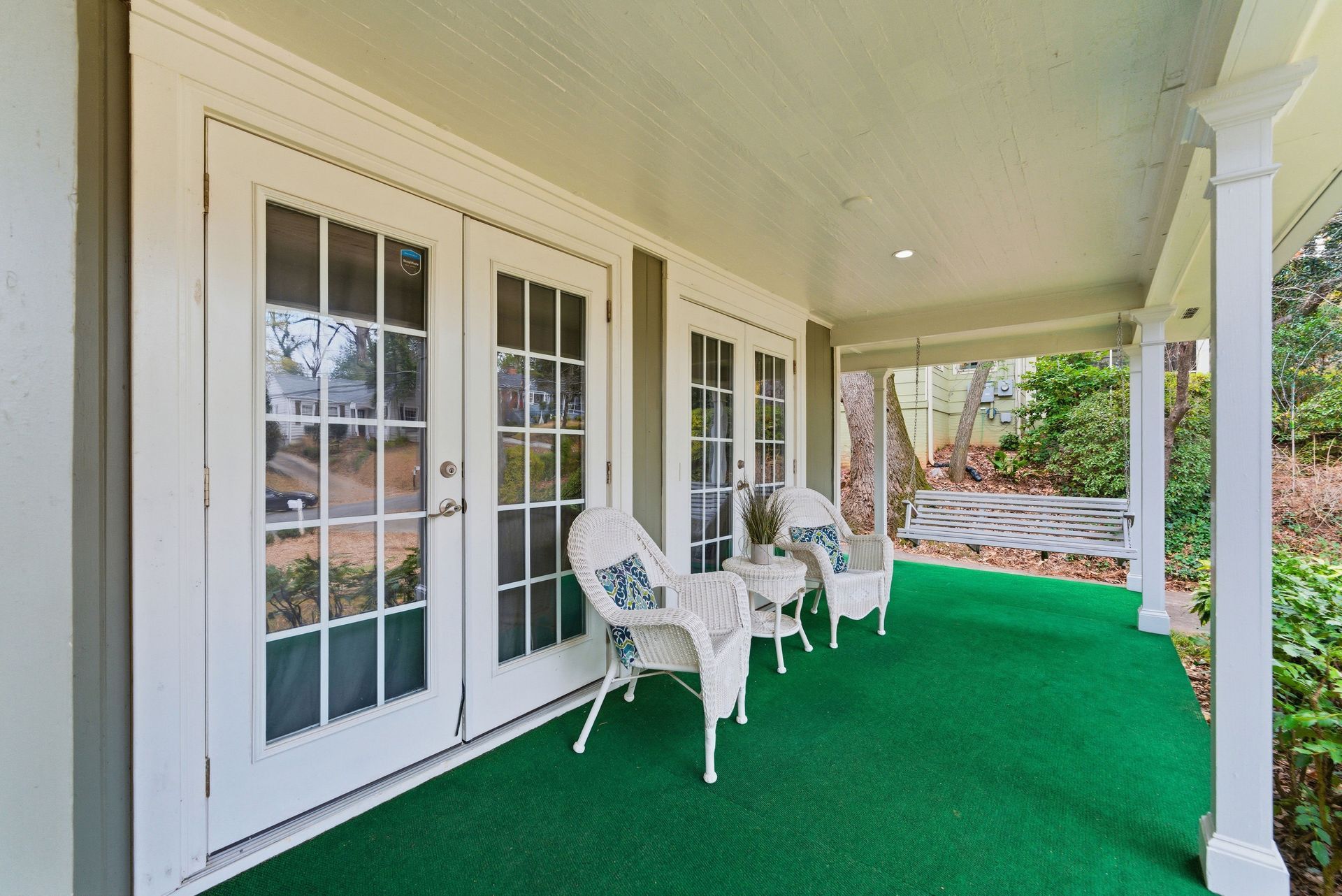 A covered porch with bright green flooring, two white wicker chairs, and a hanging porch swing by white French doors.