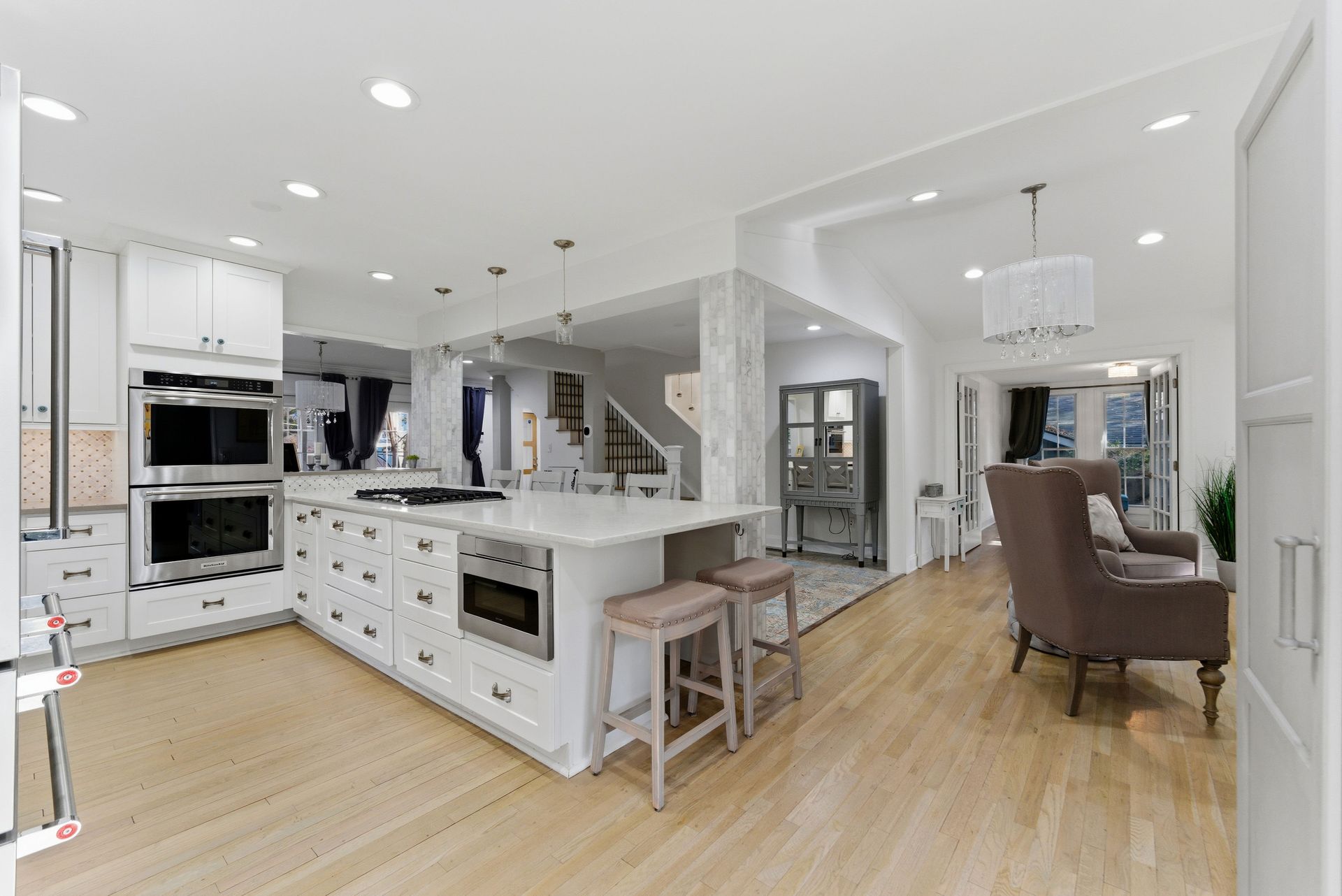 A bright, white kitchen with a large island, stainless steel appliances, and wood flooring leading to a living area.