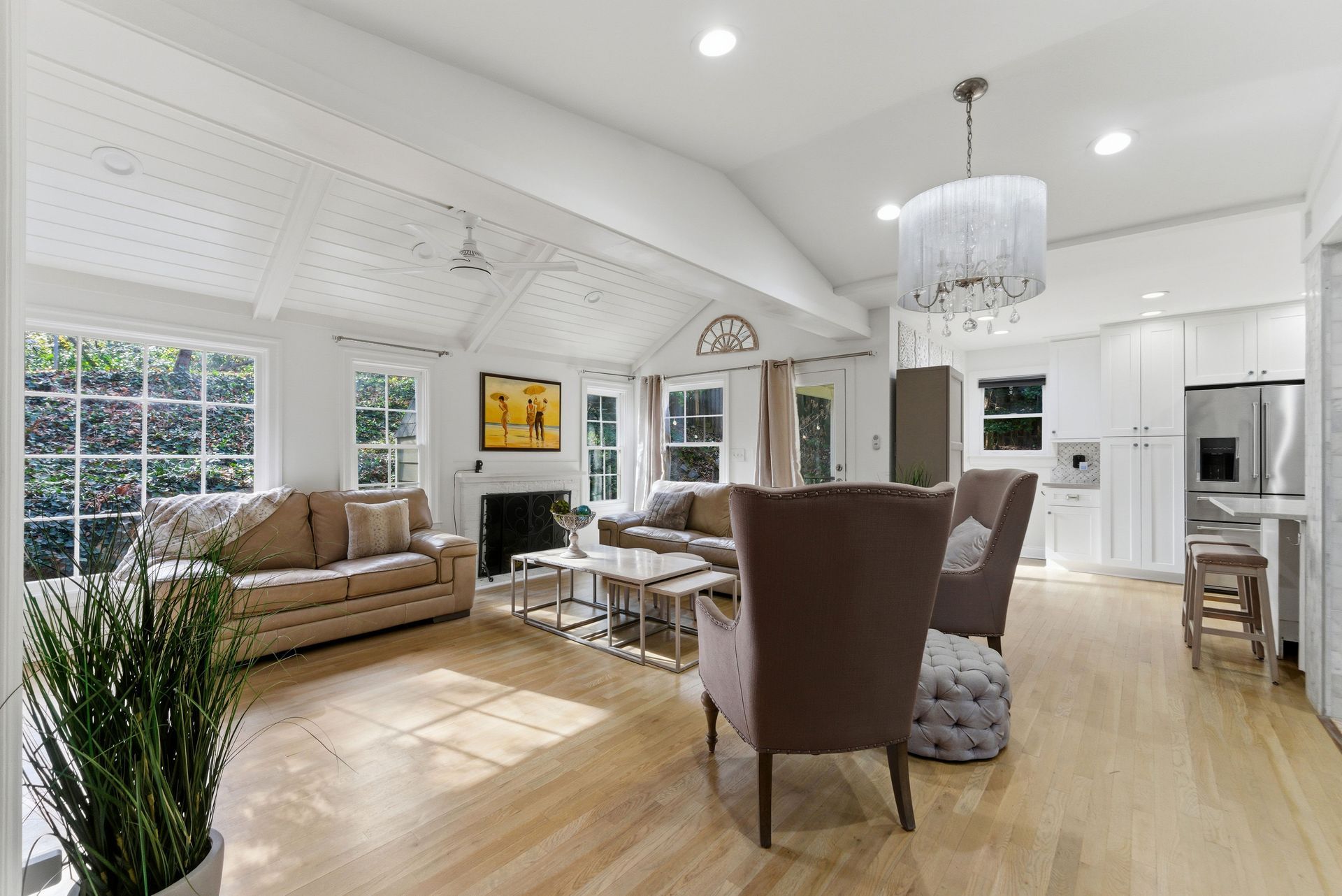 A bright, open-plan living room with vaulted ceilings, beige sofas, armchairs, hardwood floors, and a kitchen area.