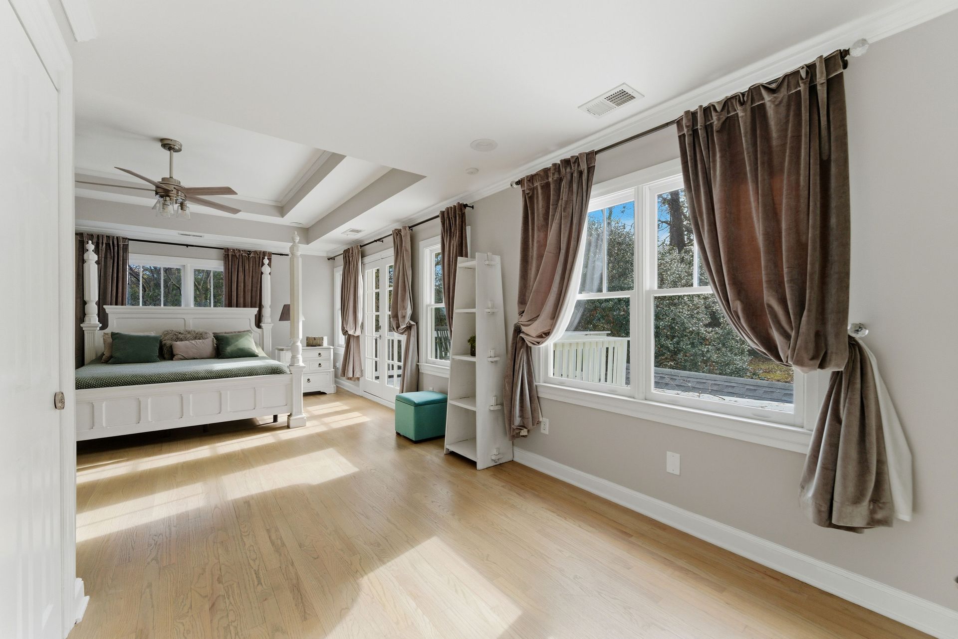 A spacious bedroom featuring a white bed with green bedding, light wood floors, and large windows with brown curtains.
