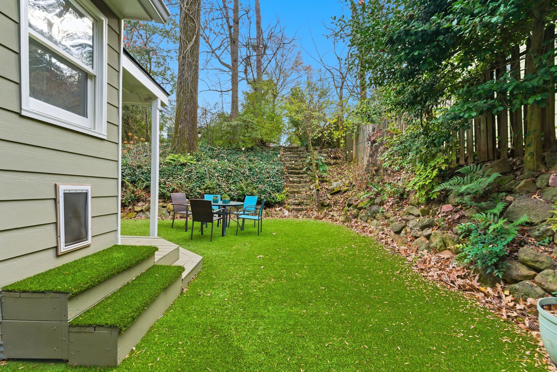 A light-green house exterior with steps covered in artificial grass leads to a backyard with seating and a rocky hillside.