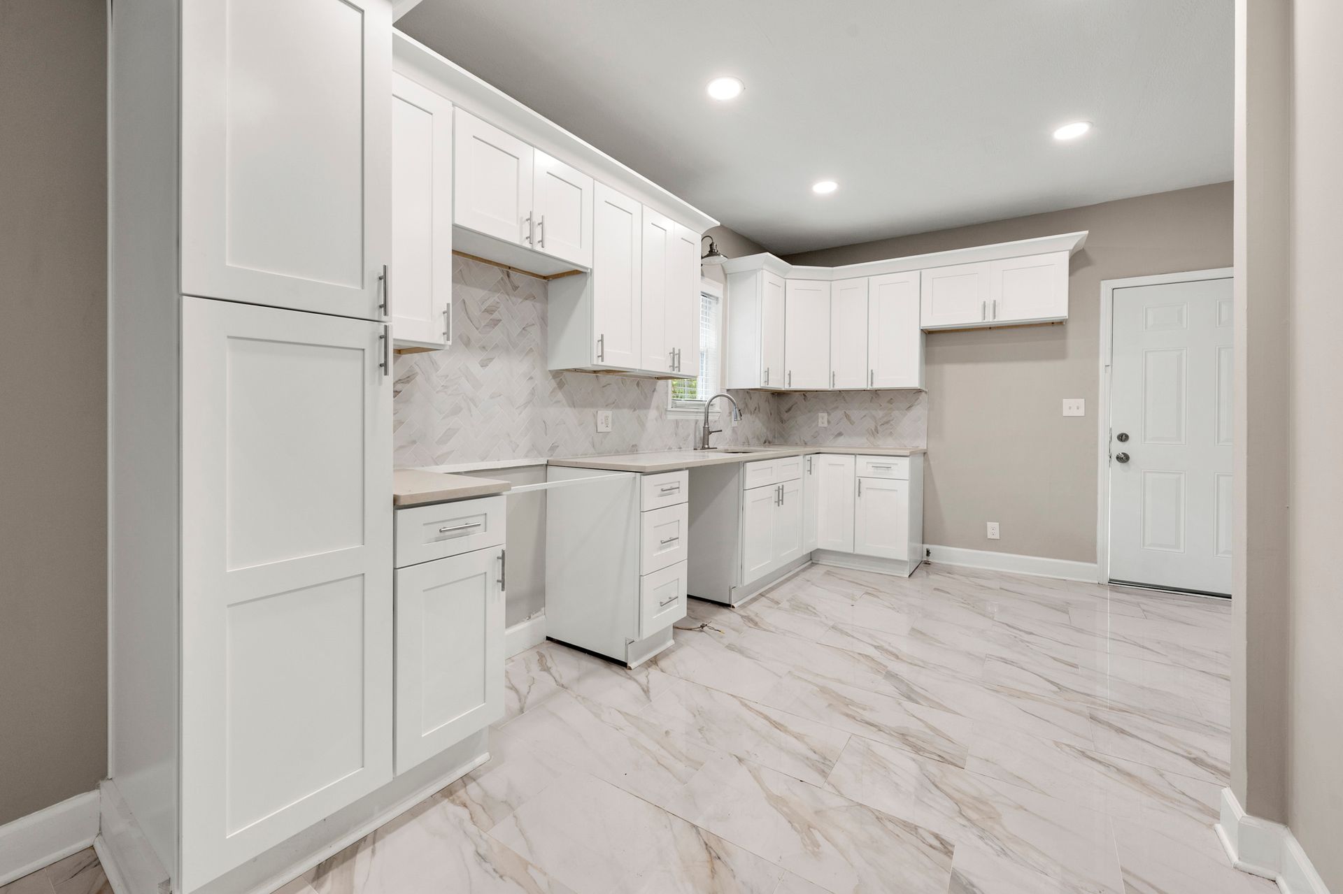 White kitchen with white cabinets, marble-look backsplash, and light-colored flooring. Empty room with a door.