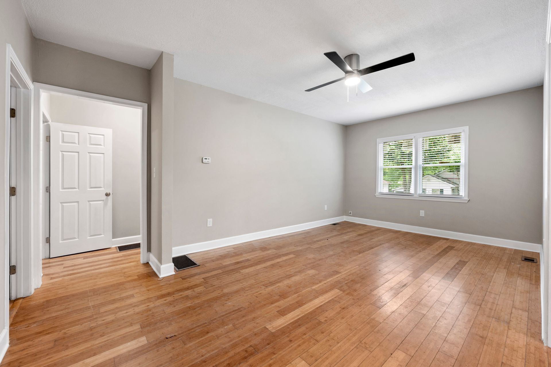 Empty room with wood floor, white trim, grey walls, doorway to a white door, window with trees, and ceiling fan.