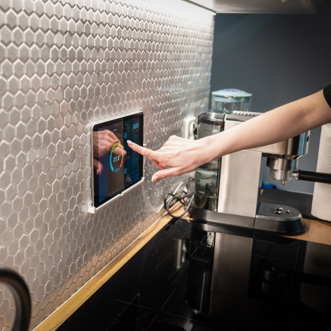 A hand touches a screen embedded in a metallic tiled kitchen wall. The screen displays a control interface.