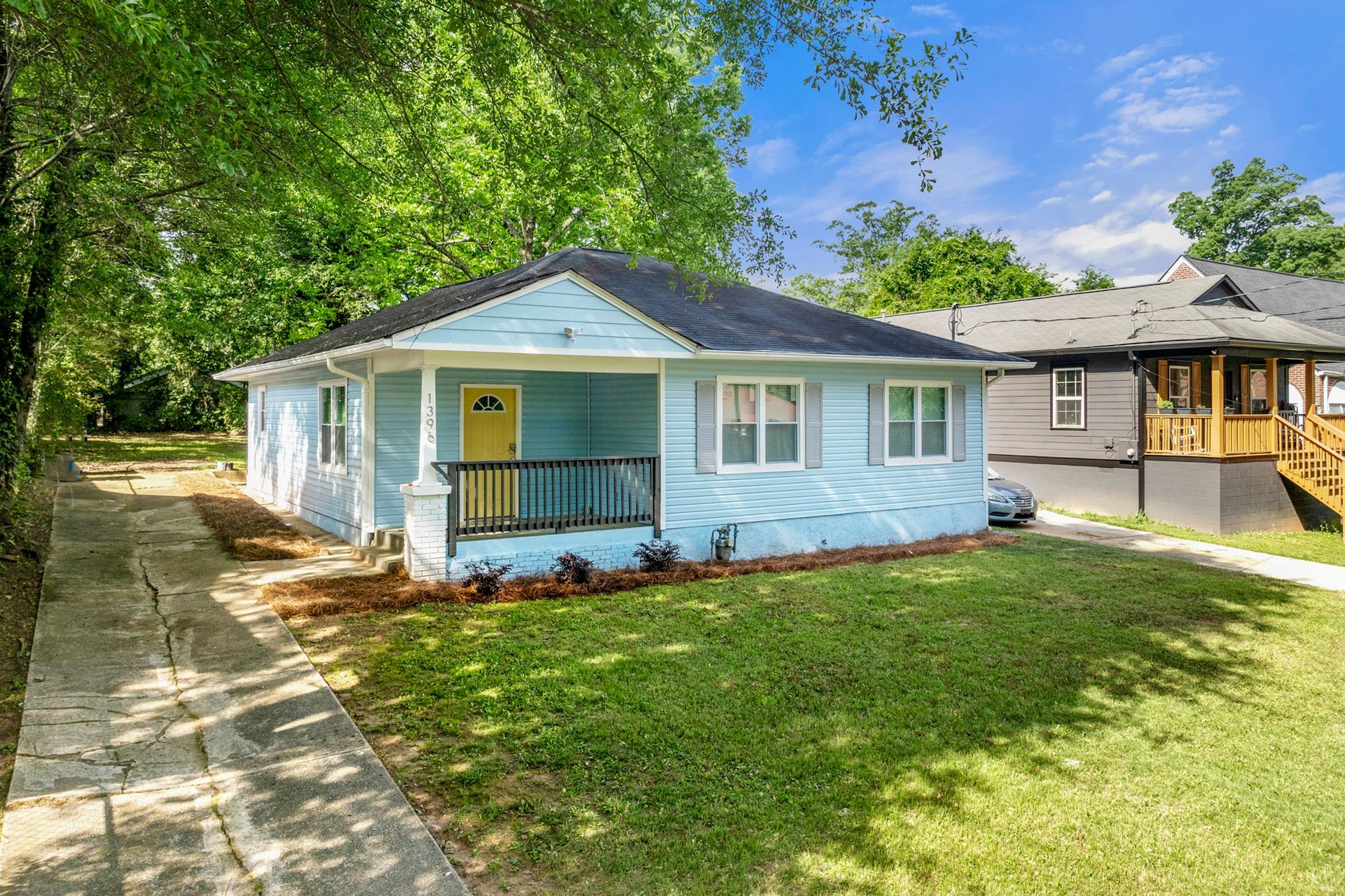 Light blue house with yellow door and black porch railing. Green lawn, trees, and driveway.