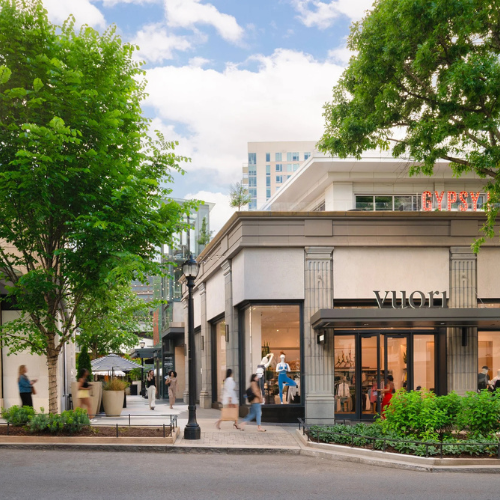 Exterior of a retail store with window displays, sidewalk, trees, pedestrians, and surrounding buildings.