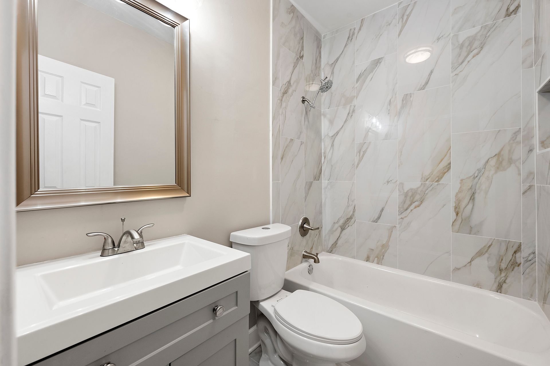 Bathroom with white sink and toilet, gray vanity, marble shower tiles, and framed mirror.