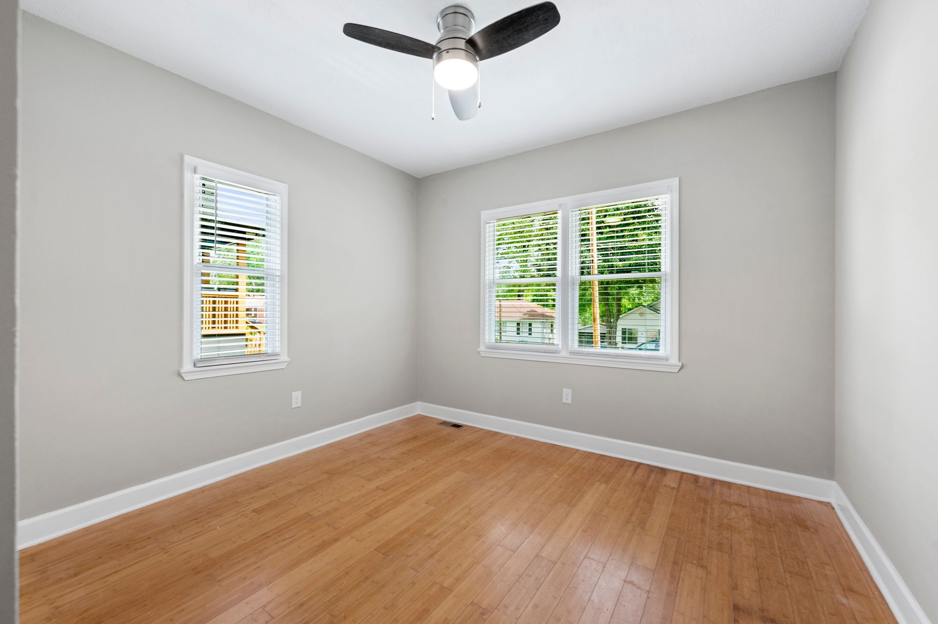Empty bedroom with hardwood floors, two windows, and a ceiling fan; light gray walls and trim.