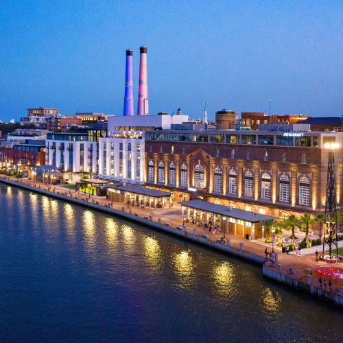 A waterfront hotel and industrial building at dusk, featuring two illuminated smokestacks and reflections in the water.