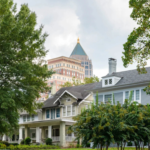 A historic pale-colored house with a porch sits in the foreground, with a tall, pointed skyscraper in the background.