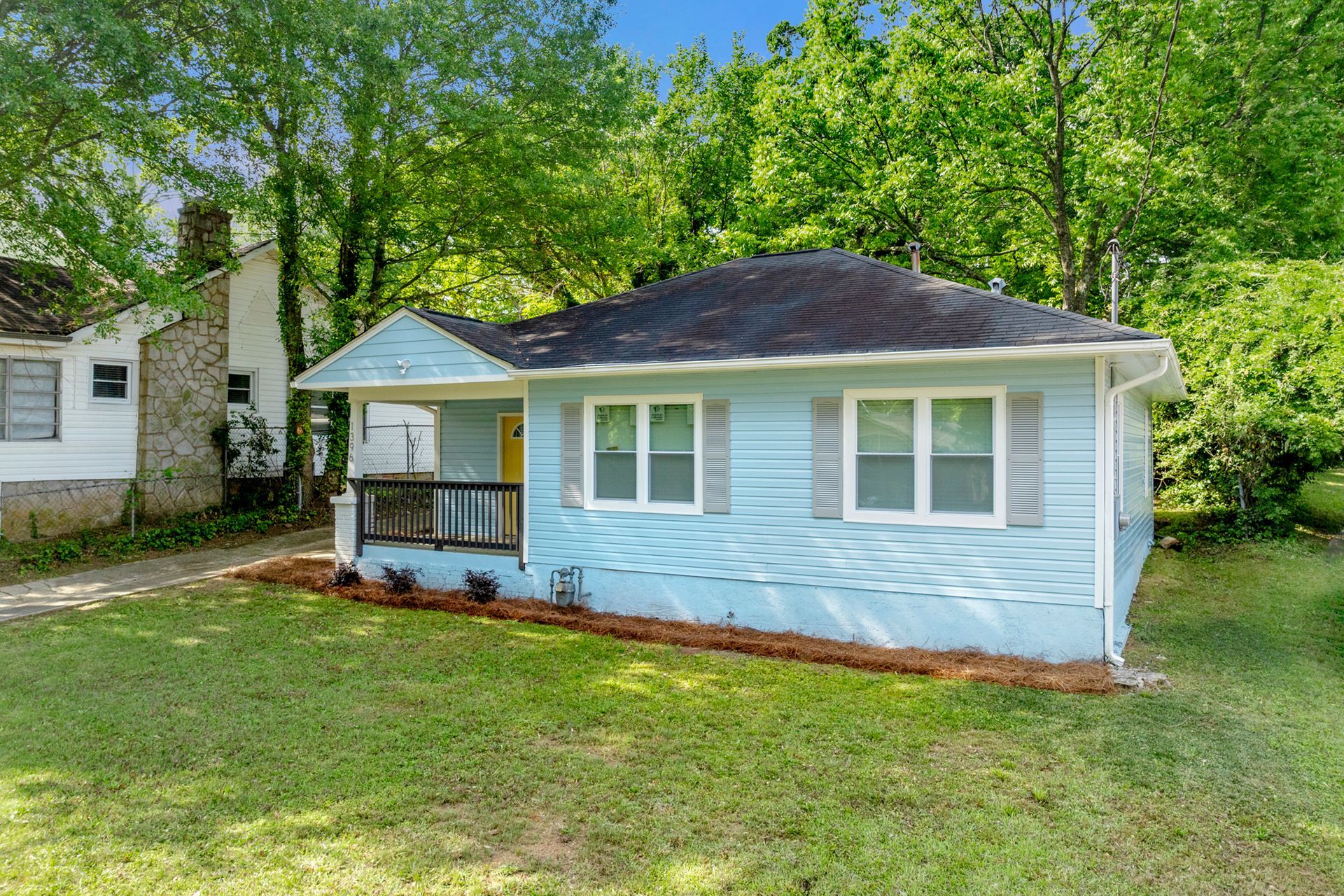 Blue bungalow with white windows and shutters, yellow door, surrounded by green grass and trees.