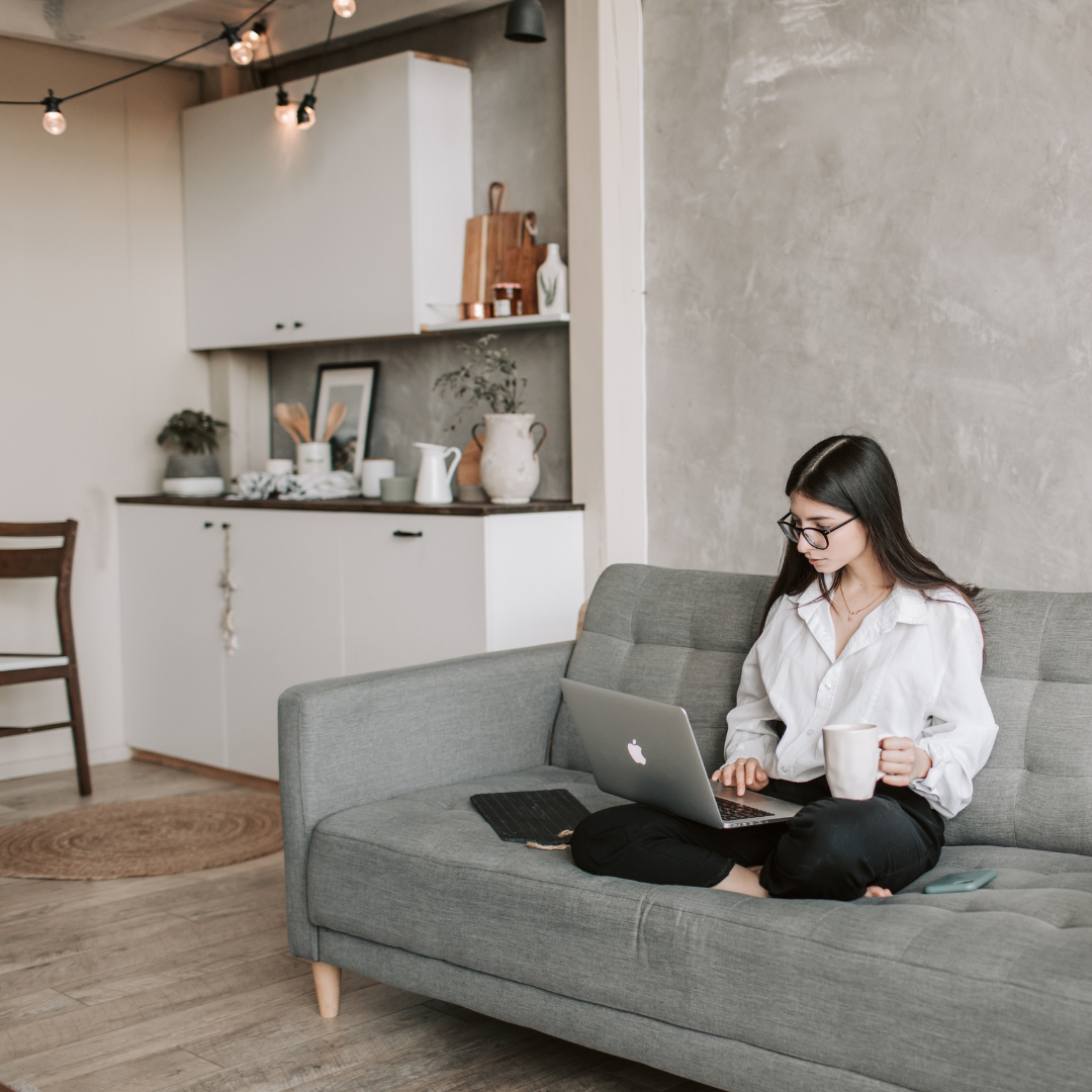 Woman on a gray sofa using a laptop, holding a mug. The setting is a living room with a kitchen in the background.