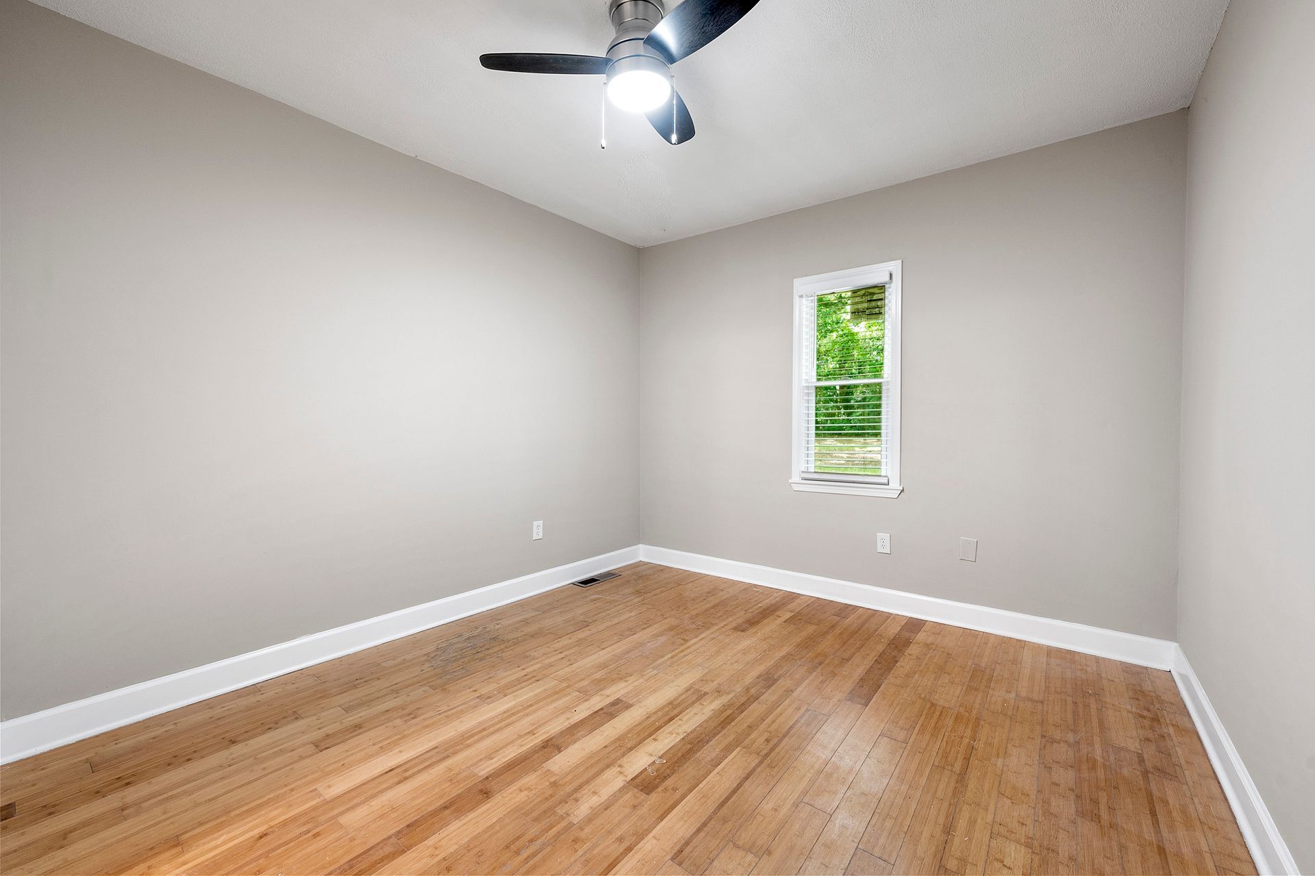 Empty room with hardwood floors, a window, and a ceiling fan; light gray walls and white trim.